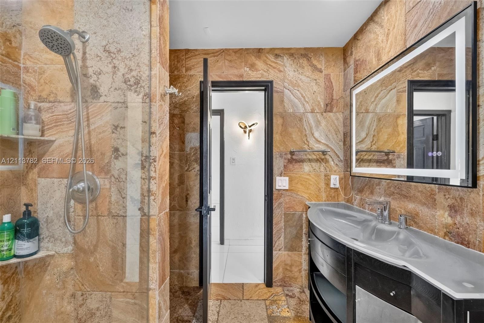 Bathroom featuring natural stone tile wall and floor surfaces, a curved vanity with a basin sink, an integrated-lighting rectangular mirror, and a wal