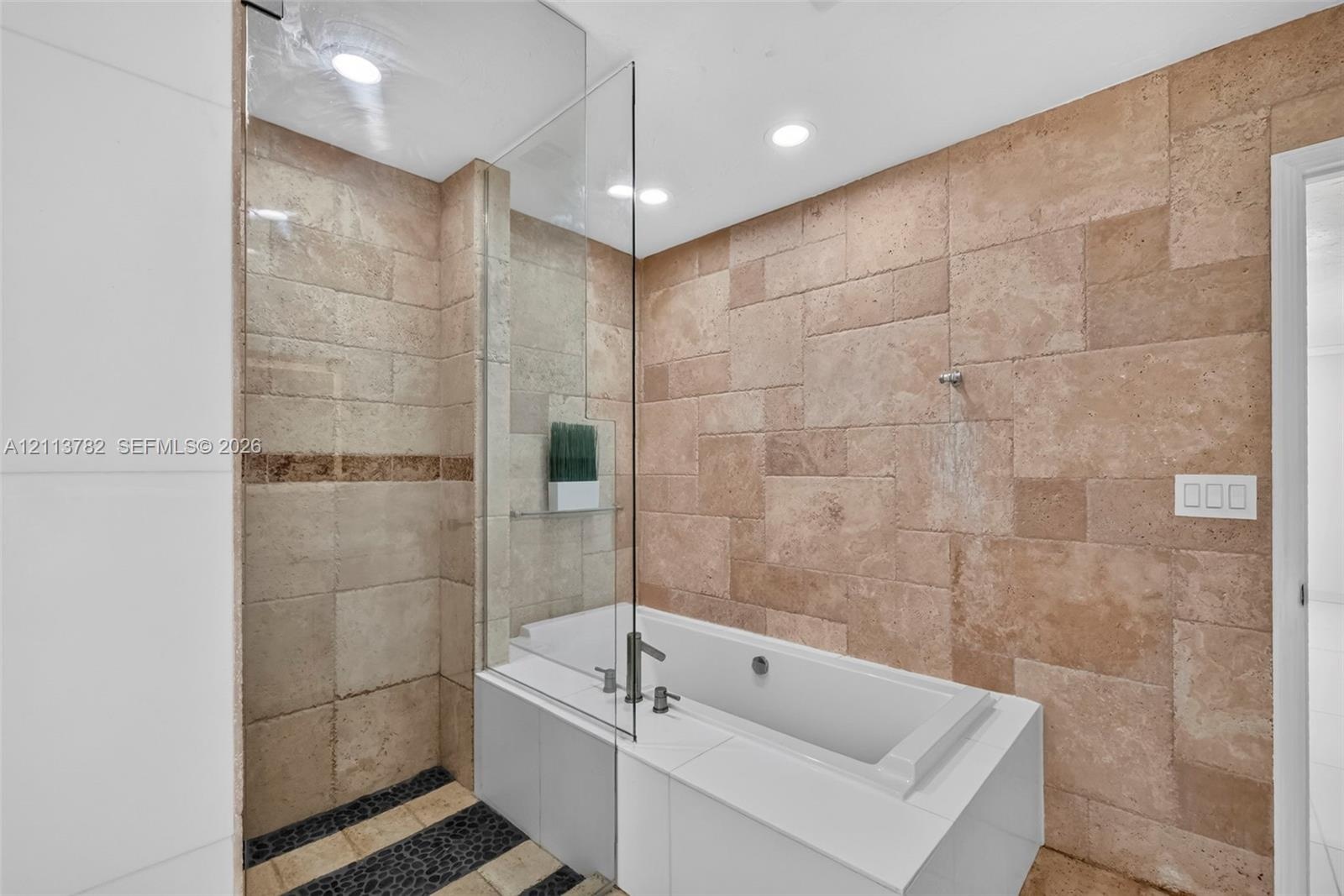 Large-format natural stone wall tiling, integrated soaking tub with a white surround, glass shower partition, recessed ceiling lighting, and pebble-st