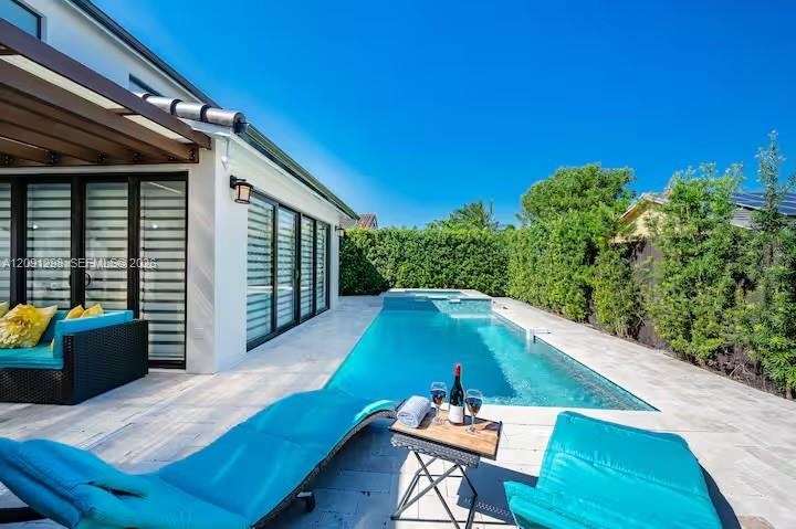 Rear exterior featuring a covered patio, rectangular in-ground pool with integrated spa, light-toned stone decking, and mature privacy hedges