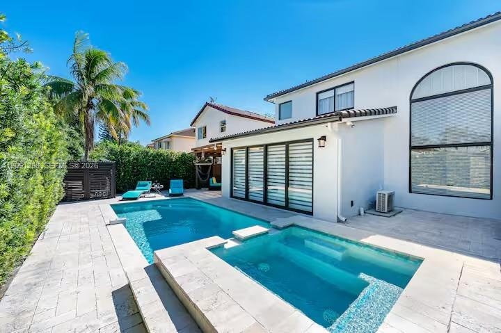 Stucco exterior featuring a multi-panel sliding glass door, arched window, integrated swimming pool with an attached spillover spa, and stone-paver ha