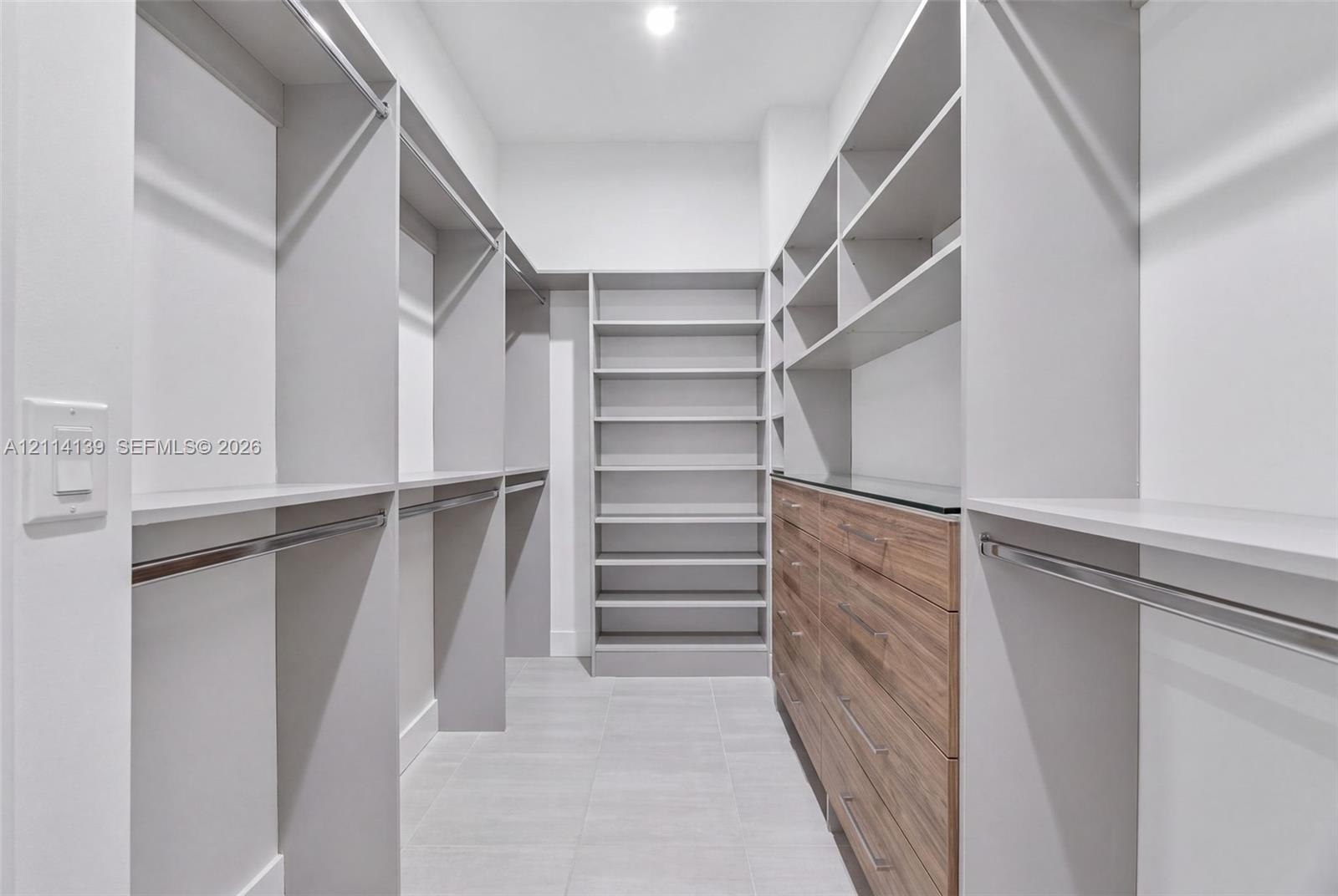 Walk-in closet featuring custom built-in shelving, wood-finish drawer cabinetry, metal hanging rods, and tiled flooring