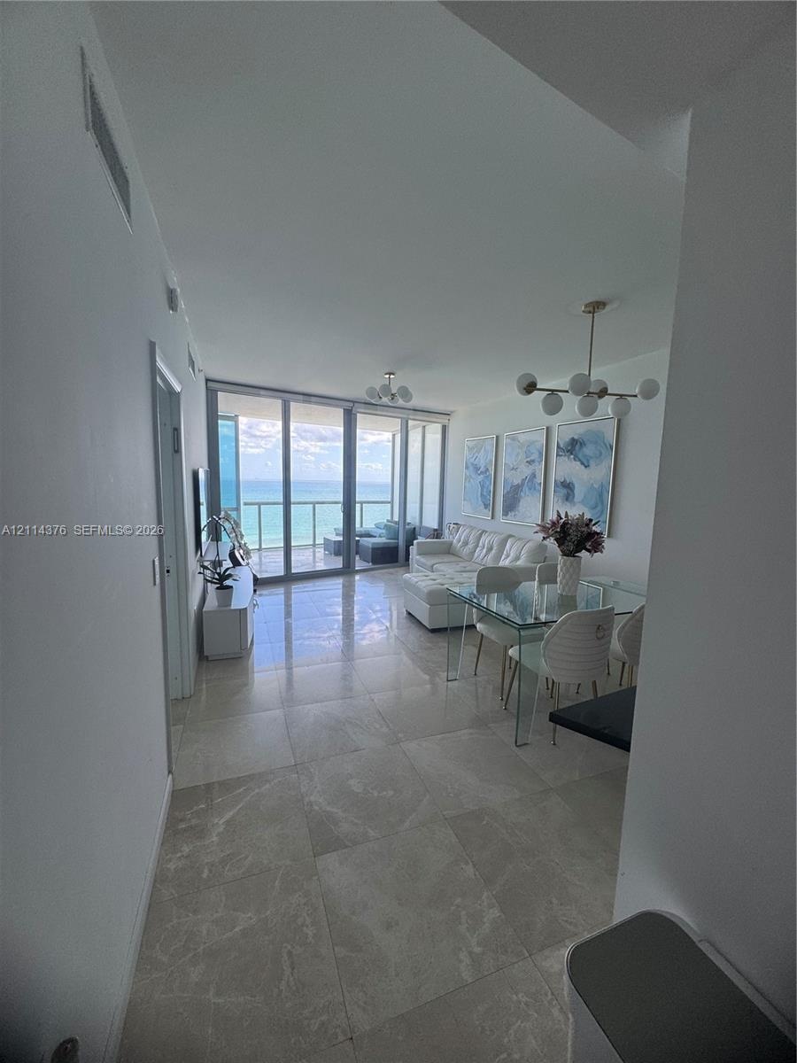 Large-format tile flooring, multi-globe metallic chandelier, floor-to-ceiling glass paneling, and an external balcony featuring a glass railing with a