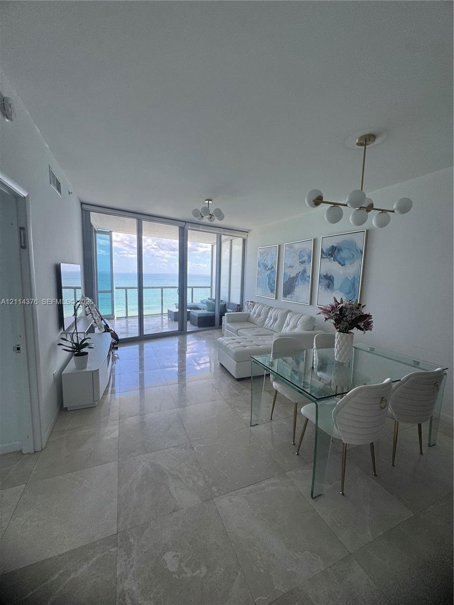 Open-concept living space featuring floor-to-ceiling glass panels, large-format tile flooring, multi-globe brass-finish chandeliers, and a private bal