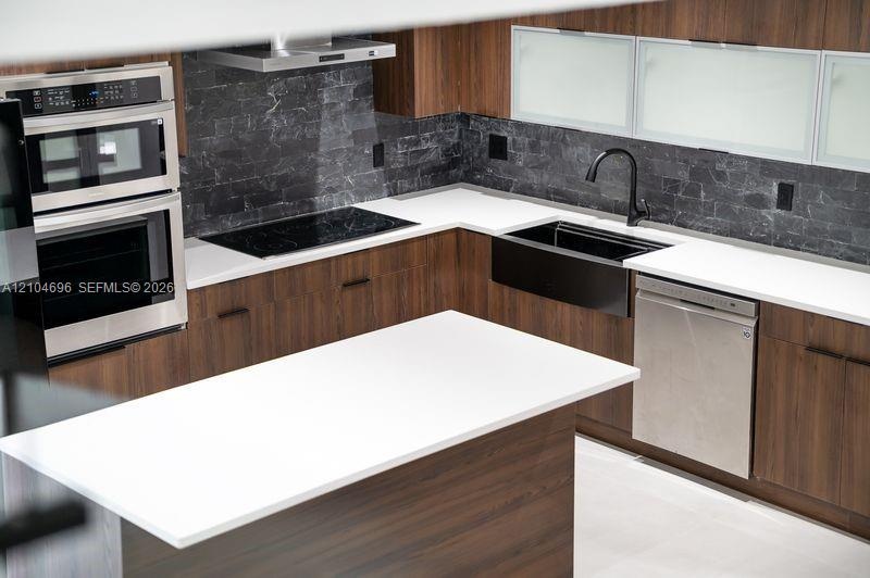 Kitchen featuring wood-finish cabinetry, white countertops, a dark stone backsplash, a black farmhouse-style sink with a matte black faucet, and stain