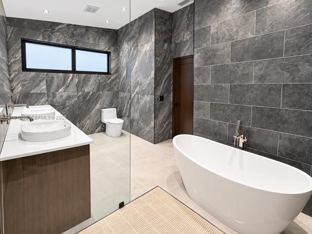 Freestanding white soaking tub with wall-mounted polished chrome faucet, dual vessel sinks atop a wood-finish vanity, and floor-to-ceiling textured st