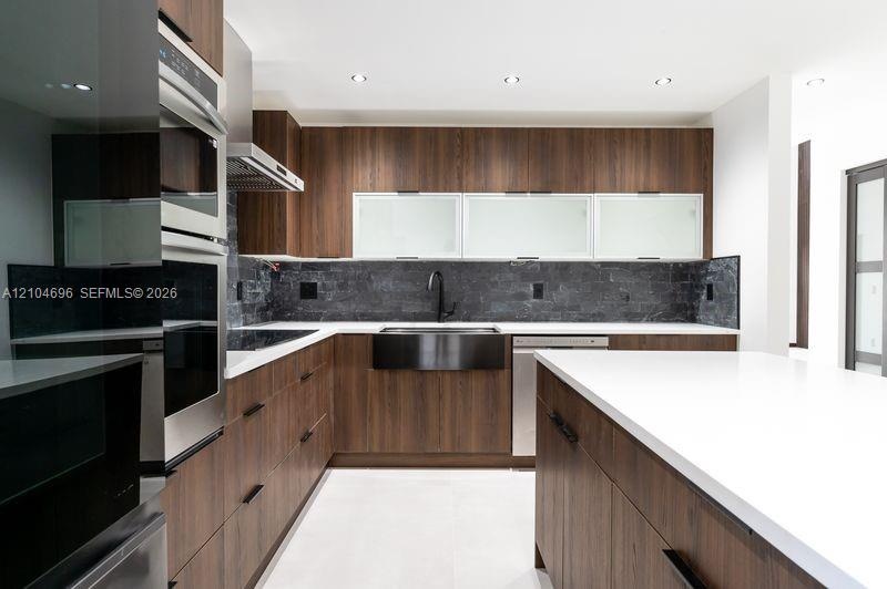 Kitchen featuring wood-finish cabinetry, white countertops, a central island, integrated stainless steel appliances, and a dark stone tile backsplash