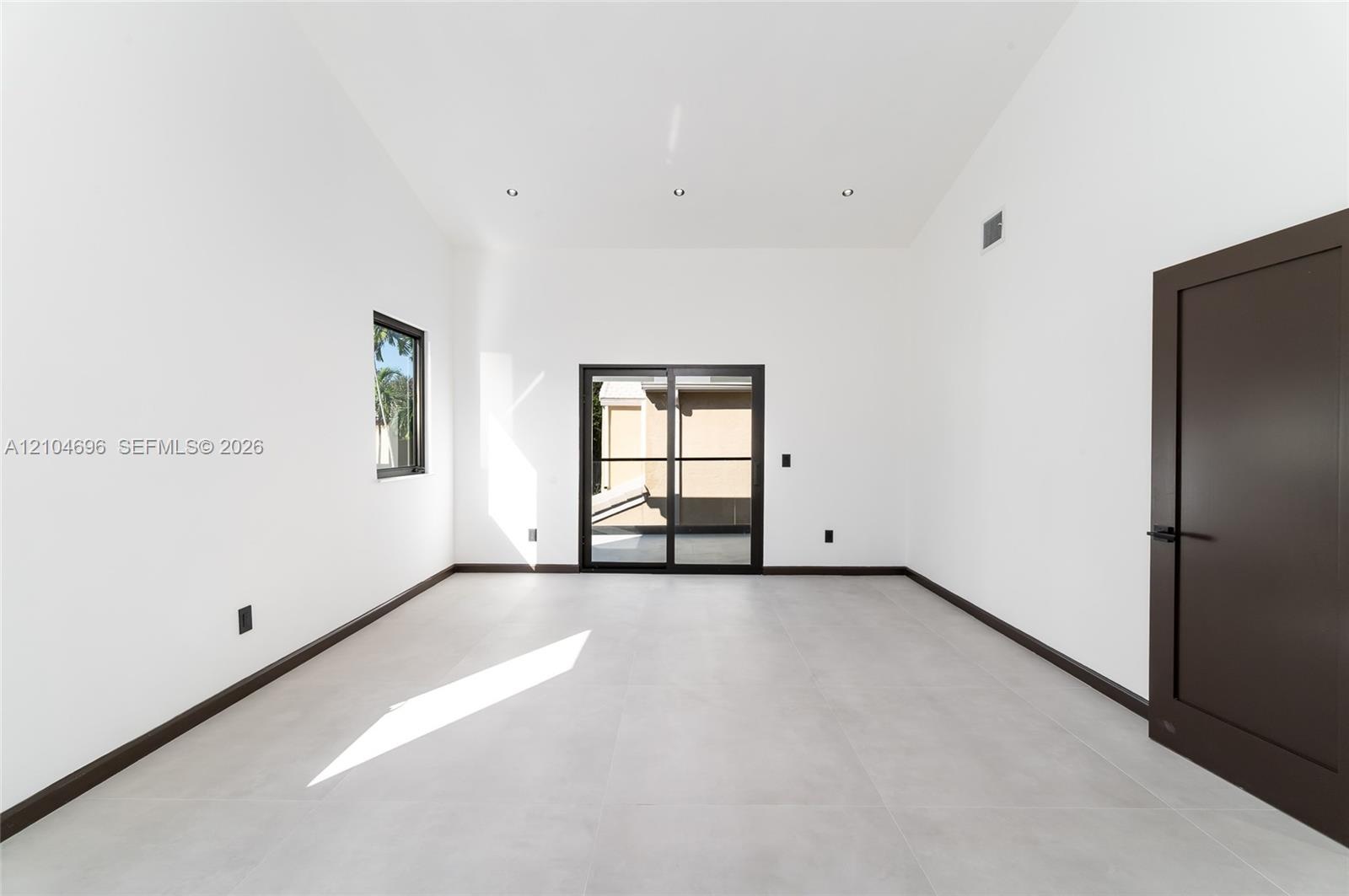 High-ceiling interior featuring large-format tile flooring, recessed lighting, dark-framed sliding glass doors, and a solid-core door with matte black