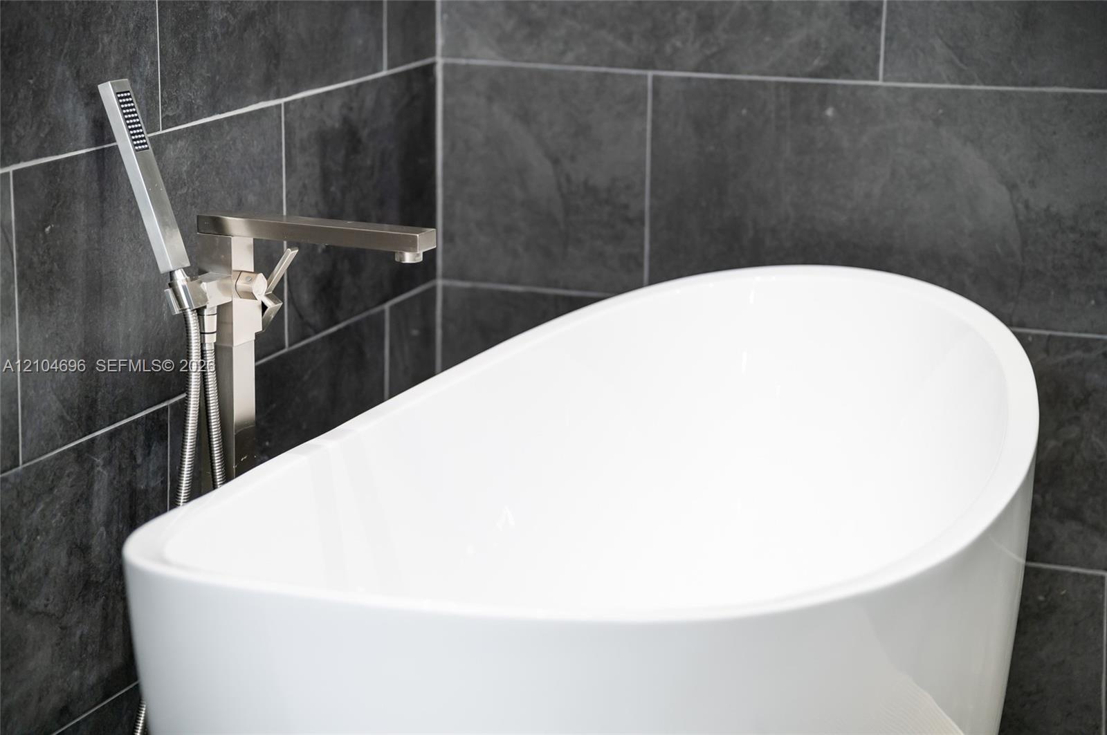 Freestanding white soaking tub featuring a metallic finish floor-mounted faucet with an integrated handheld sprayer, set against charcoal-toned wall t