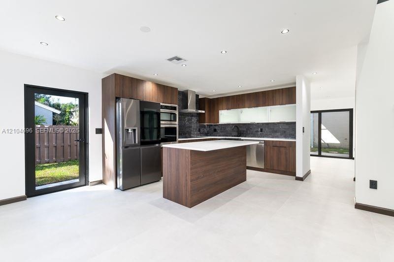 Kitchen featuring flat-panel wood-finish cabinetry, a central island with a solid-surface countertop, integrated stainless steel appliances, dark ston