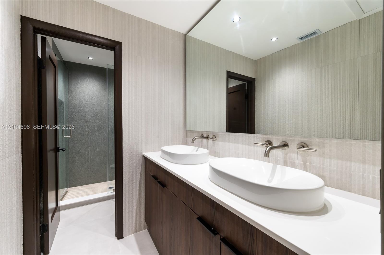 Floating vanity featuring a dual-sink countertop, vessel sinks, wall-mounted faucets, large frameless mirror, and a walk-in shower with glass enclosur