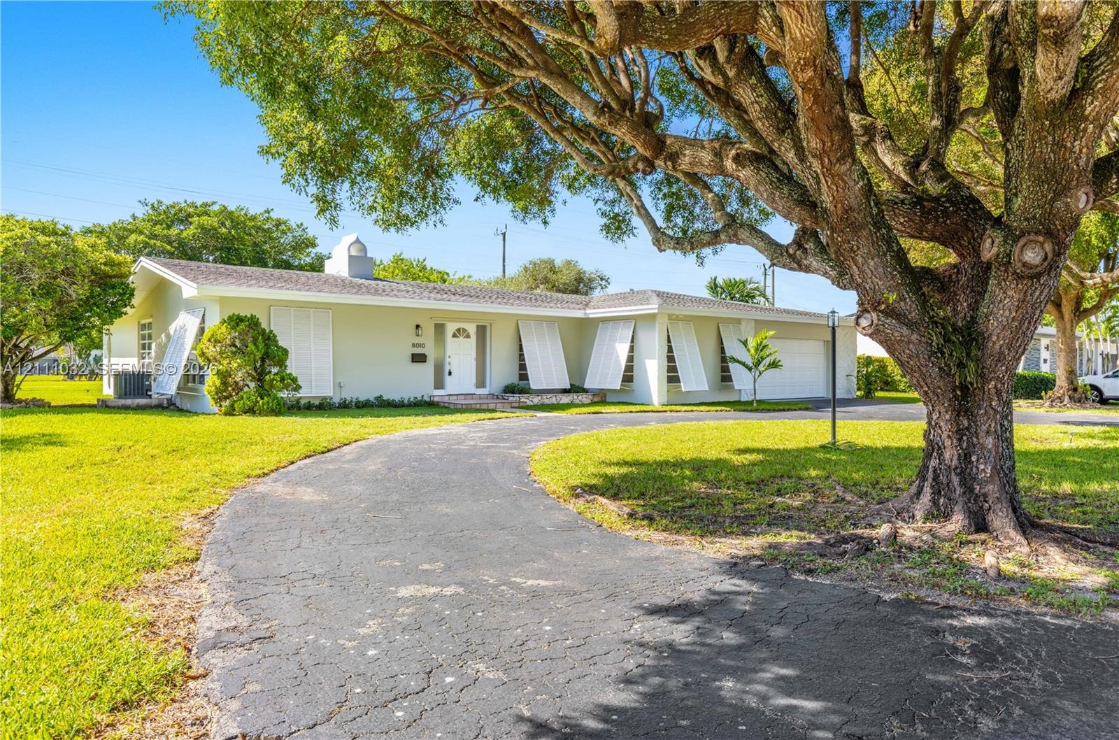 Single-story residence featuring a curved paved driveway, exterior shutters, attached garage, and mature landscaping on a spacious lot