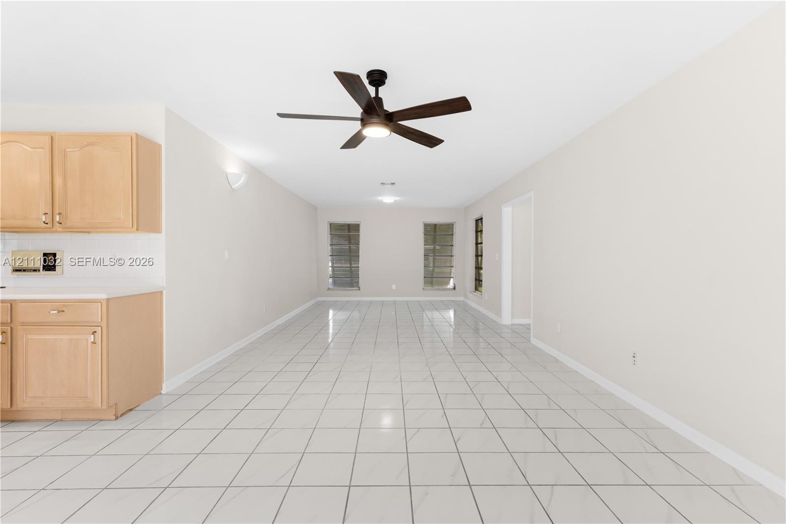 Open-concept area featuring light-tone cabinetry, ceramic tile flooring, three large windows, and a central ceiling fan with integrated lighting