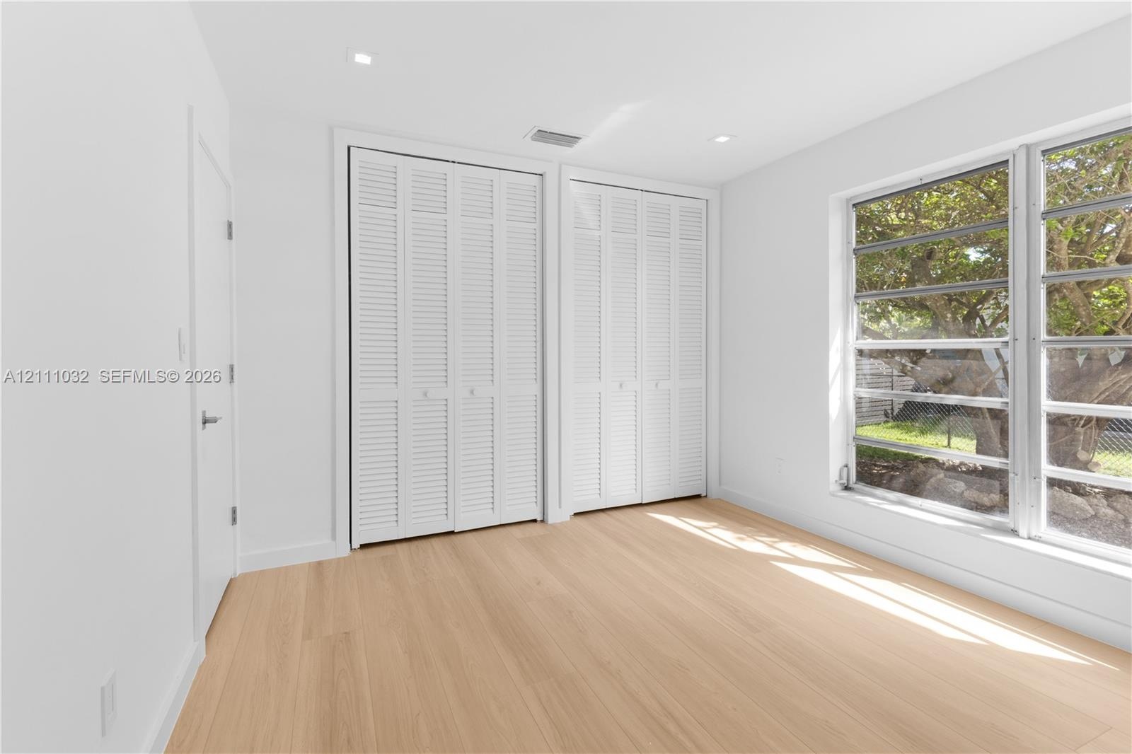 Room interior featuring wood-finish flooring, white walls, dual louvered-door closets, and multi-pane windows with exterior greenery