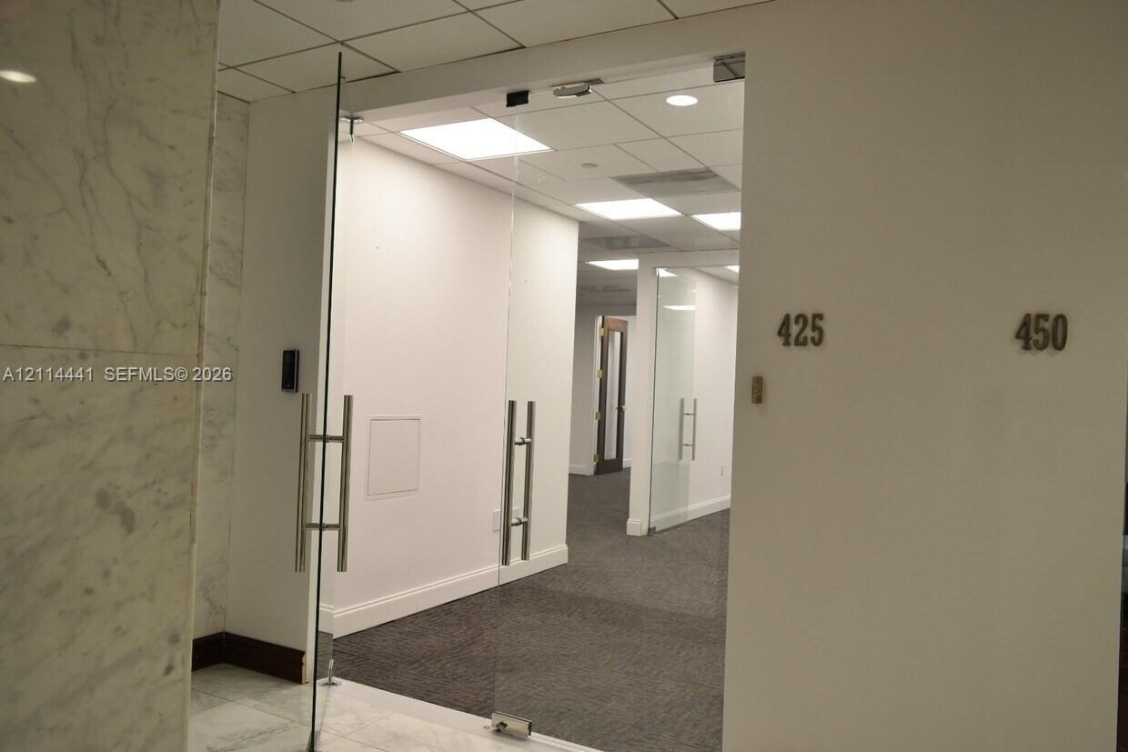 Frameless glass entry door featuring brushed metal hardware, suspended acoustic tile ceiling with recessed lighting, and stone-tiled foyer wall