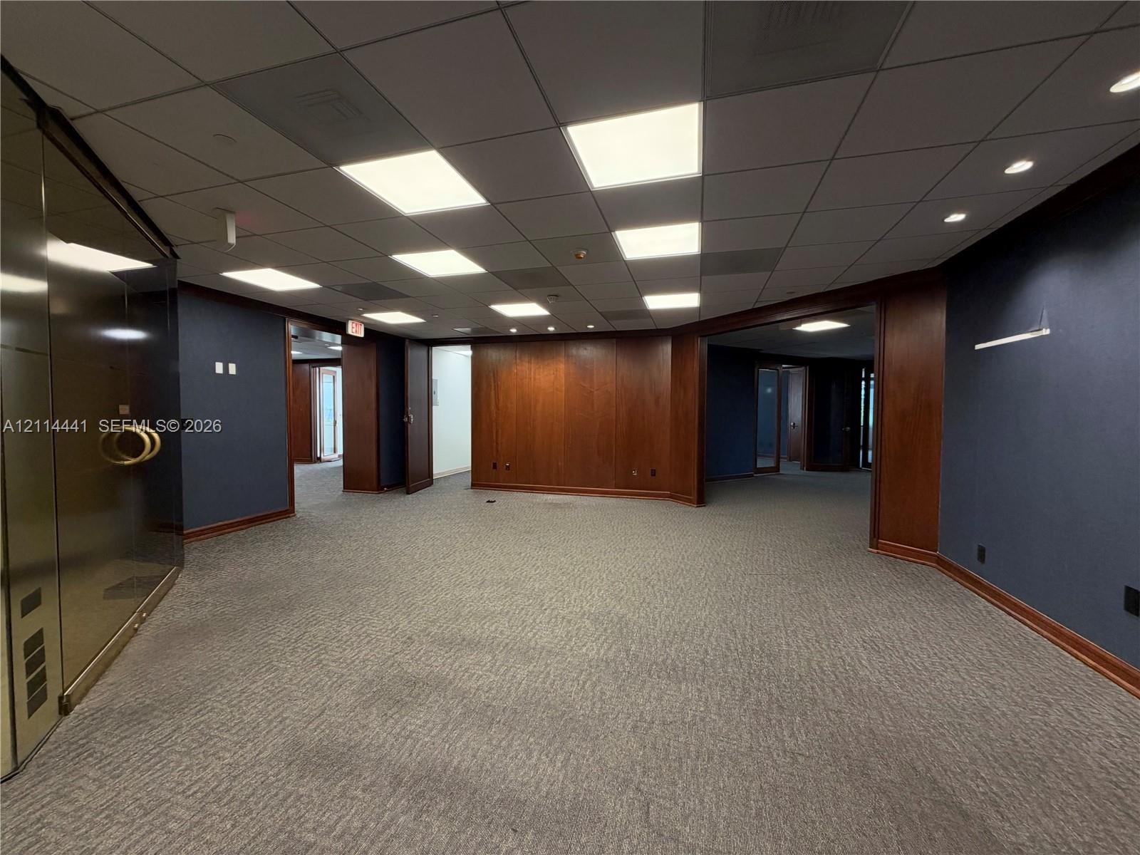 Commercial office space featuring high-contrast wood paneling, drop ceiling with recessed panel lighting, floor-to-ceiling glass partition with brass 
