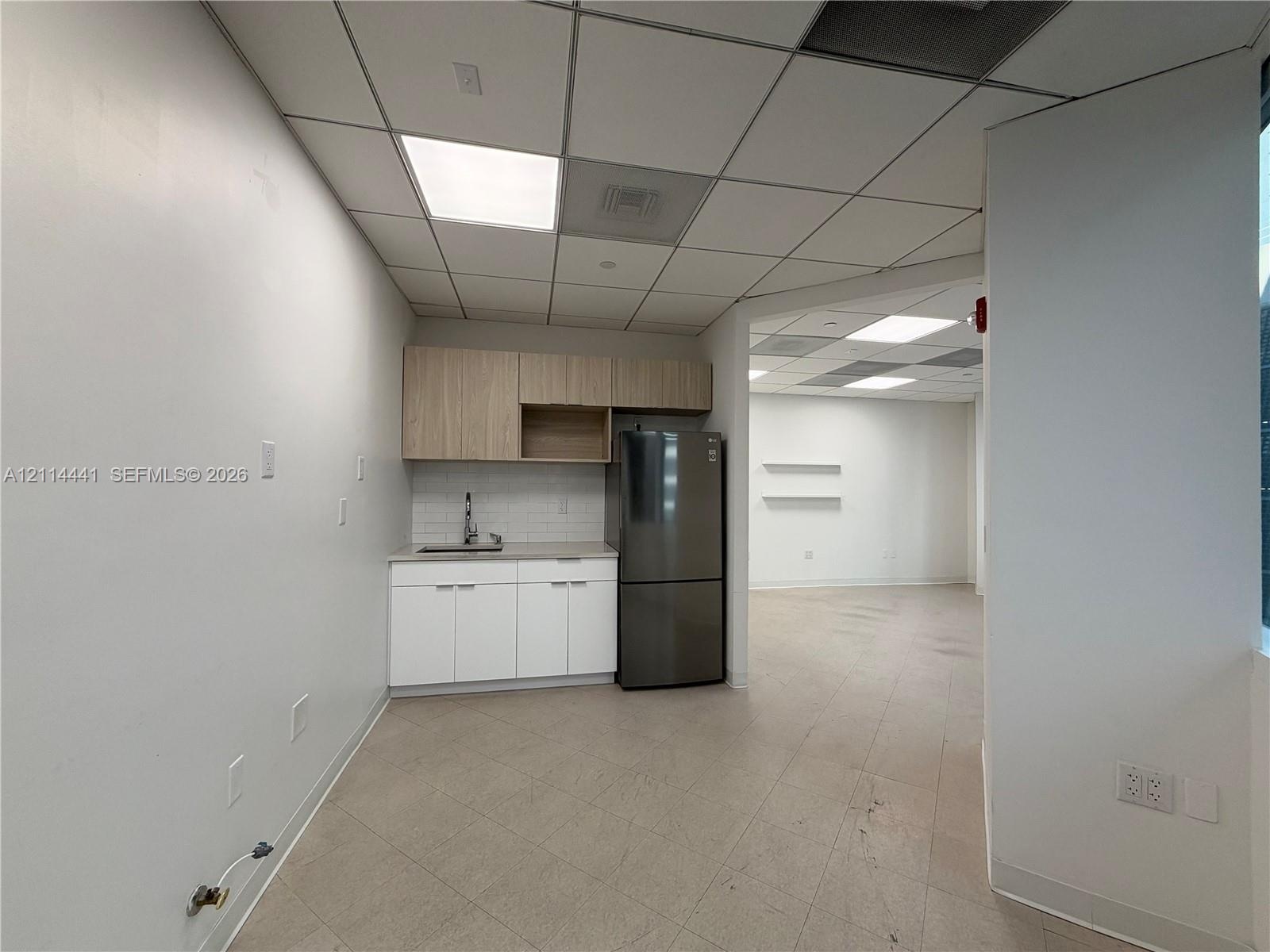 Kitchenette featuring white lower cabinetry, wood-finish upper cabinets, a white tile backsplash, a stainless steel sink with faucet, and a stainless 
