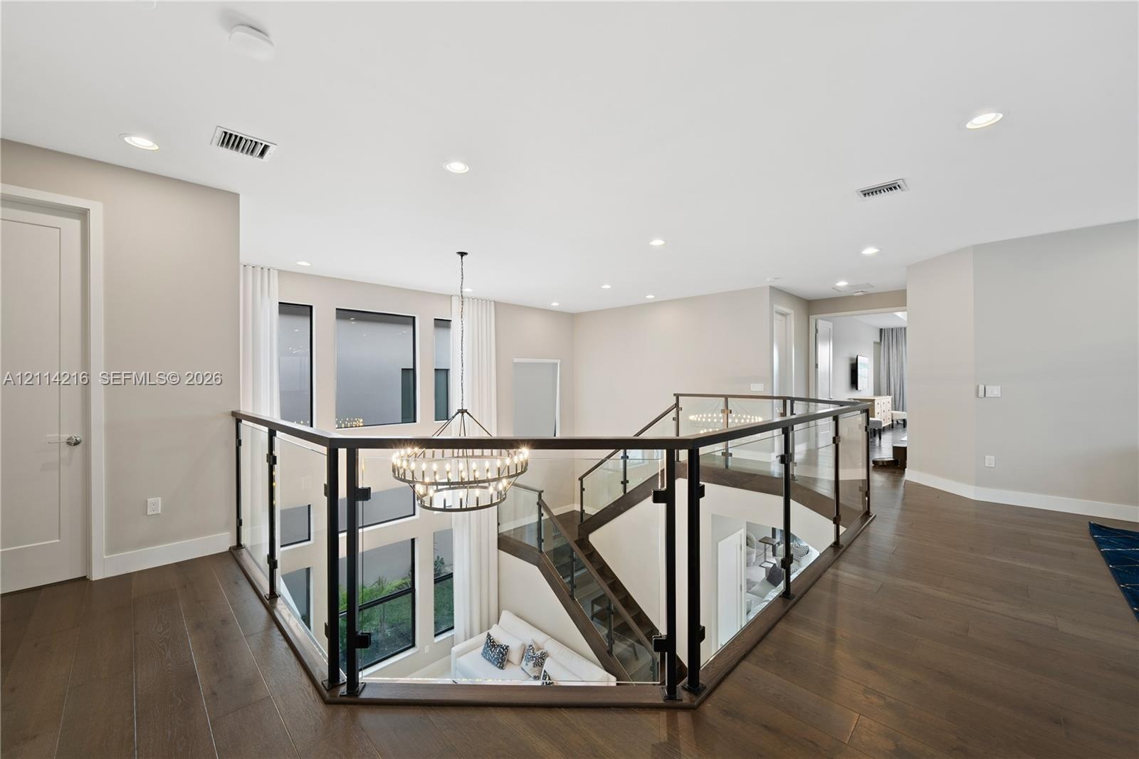 Open mezzanine landing featuring wood-finish flooring, glass panel railing with dark metal framing, recessed lighting, and a multi-tiered circular cha
