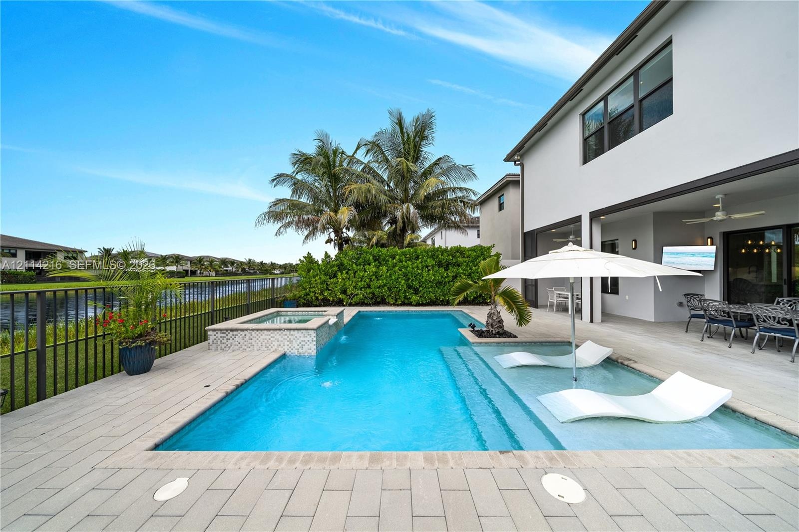 Rear patio featuring a swimming pool with a built-in spa, a tanning ledge, light-toned paver decking, and metal perimeter fencing overlooking a waterw