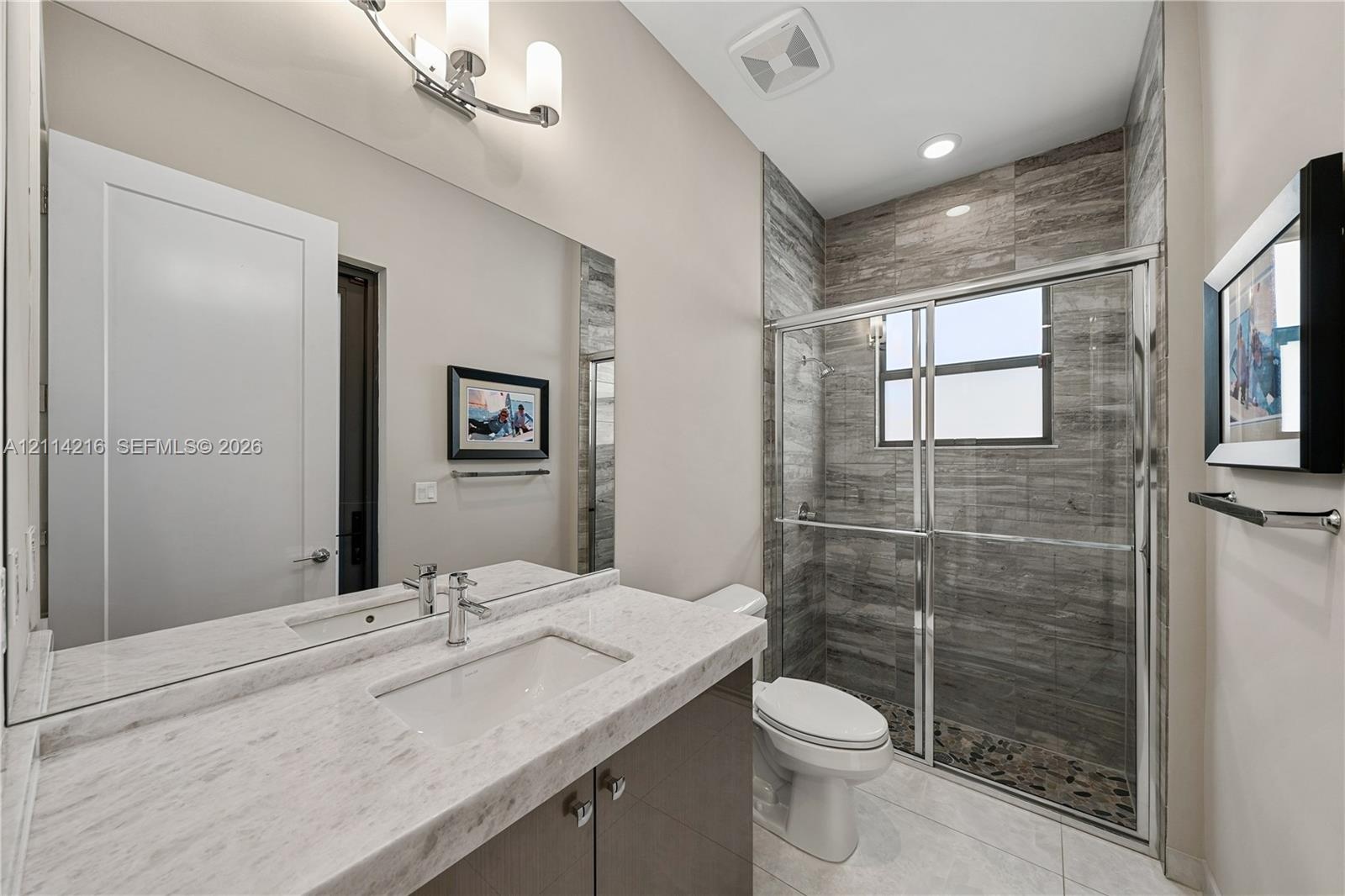 Vanity with light-toned stone-finish countertop and integrated rectangular basin, full-width mirror, glass-enclosed shower with horizontal textured ti