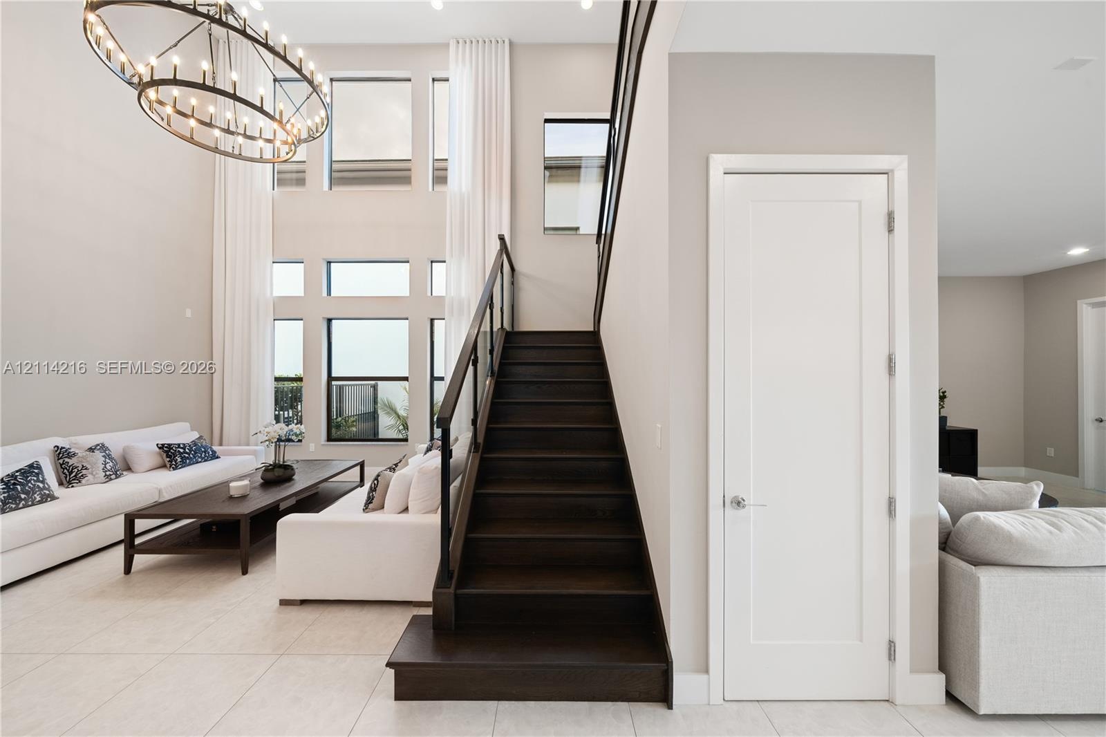 Double-height ceiling, large multi-pane windows, statement ring chandelier, staircase with wood-finish treads and a dark metal railing, and light-tone