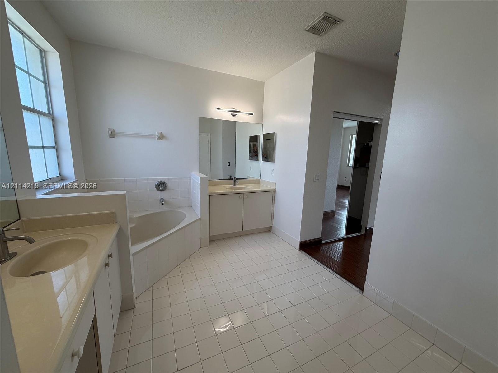 Bathroom featuring a soaking tub, dual-sink vanity with light-tone countertops, polished wall-mounted mirrors, and white ceramic tile flooring