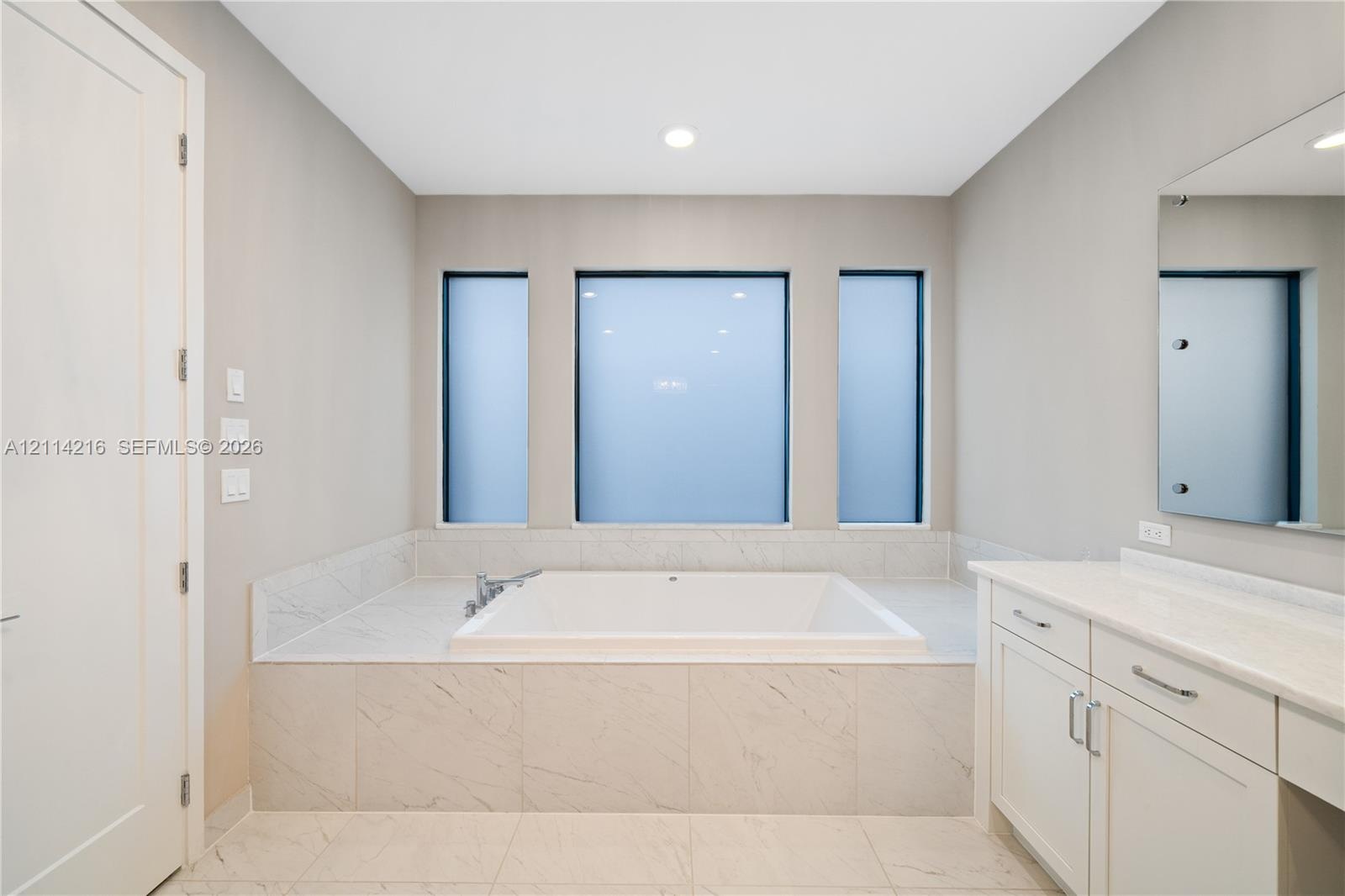 Built-in soaking tub with a tiled surround, triple-window configuration, white cabinetry with stone-finish countertops, and wall-mounted vanity mirror