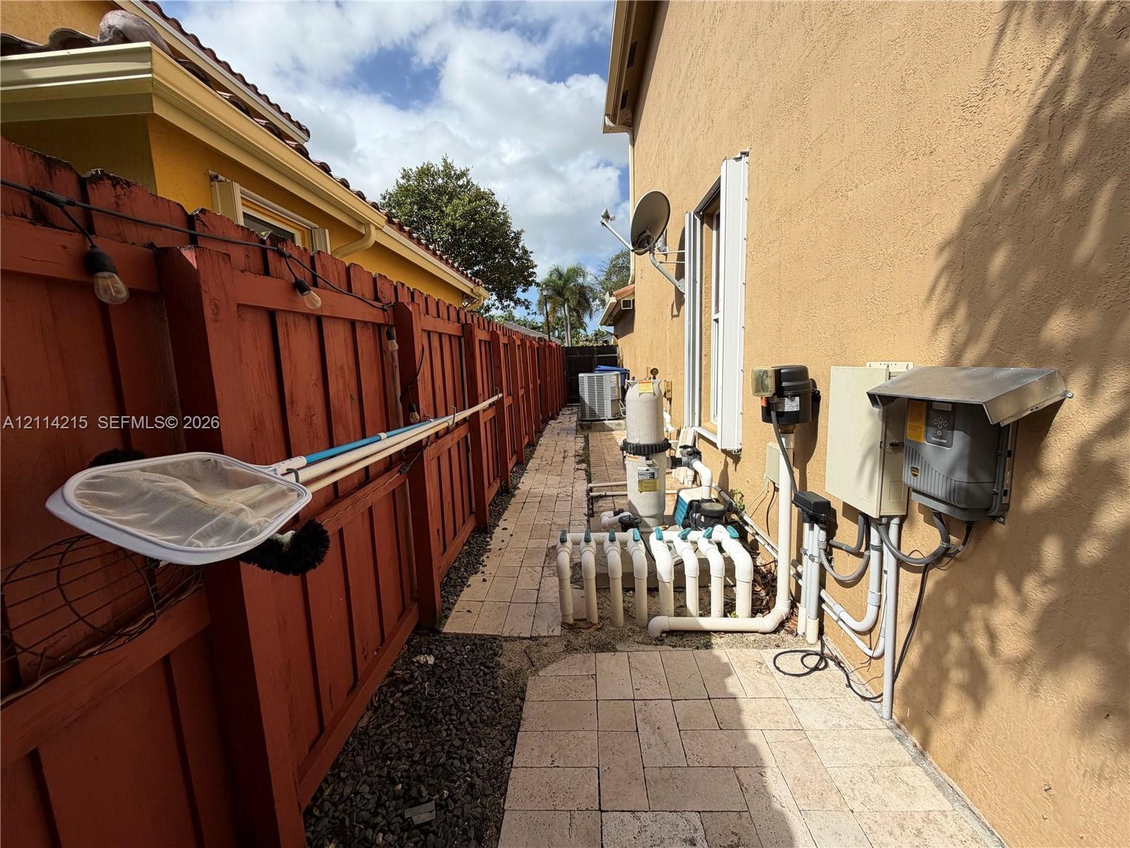 Side yard featuring paver pathways, enclosed by a wood-finish fence, with exterior-mounted pool filtration system and electrical utility equipment