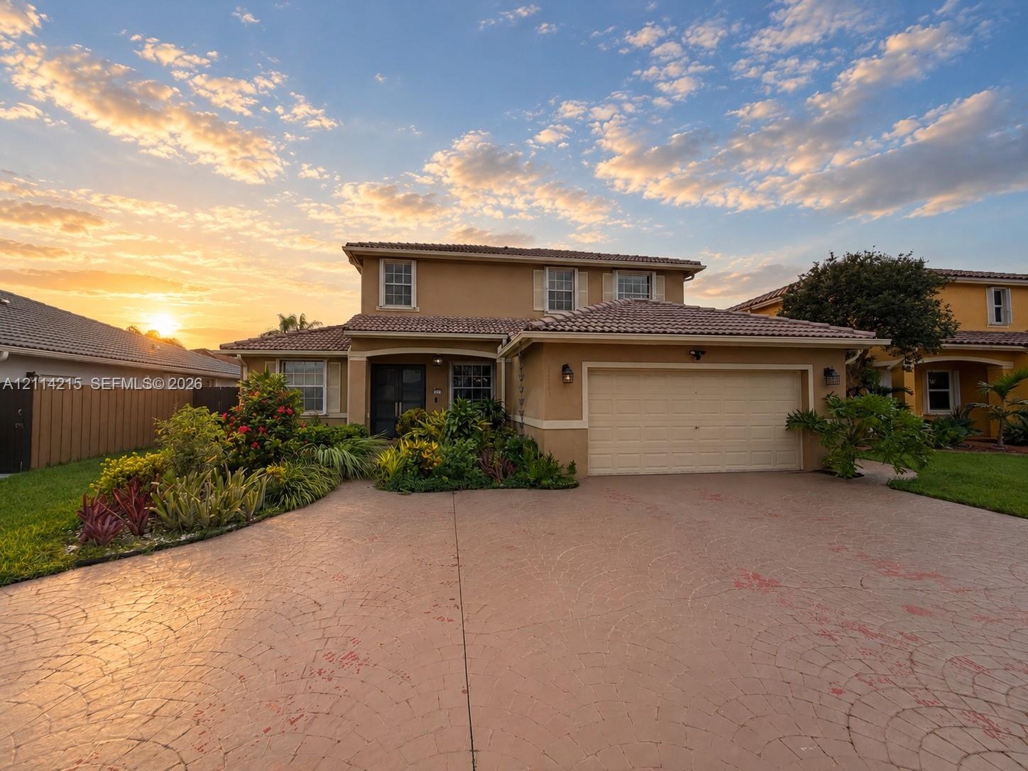 Two-story residence featuring a tile roof, stucco exterior, attached two-car garage, and a decorative stamped concrete driveway surrounded by establis
