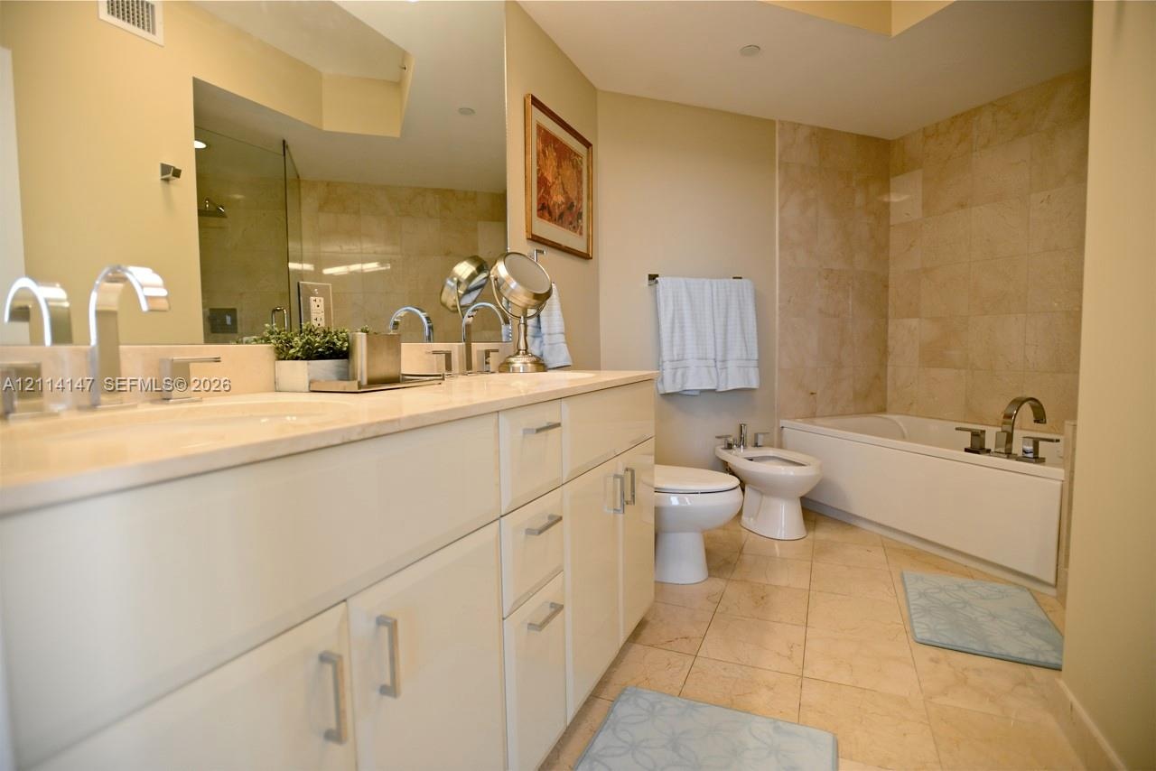 Double vanity with stone countertop, undermount sinks, and polished chrome faucets, featuring a full-width mirror, tile flooring, and a tiled shower-t