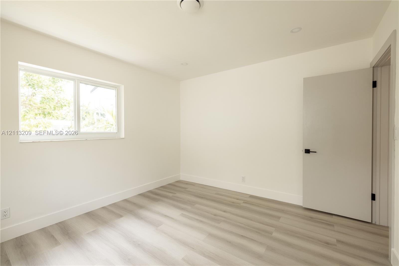Neutral-toned interior space featuring wood-finish flooring, recessed lighting, a horizontal sliding window, and a solid-panel door with matte black h