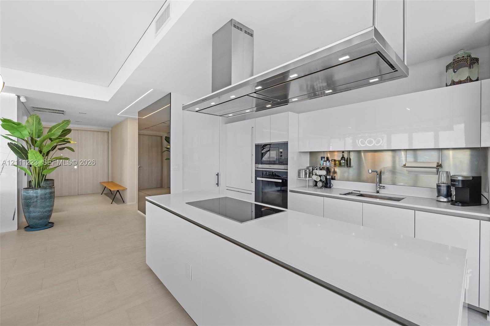 Kitchen featuring high-gloss cabinetry, a spacious island with an integrated induction cooktop, a stainless steel range hood, and a metallic backsplas