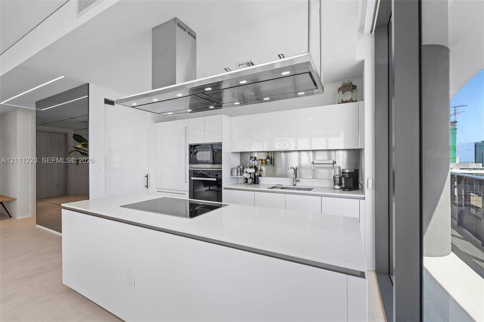 Kitchen featuring white gloss cabinetry, expansive island with integrated cooktop, stainless steel range hood, built-in wall oven, and metallic backsp