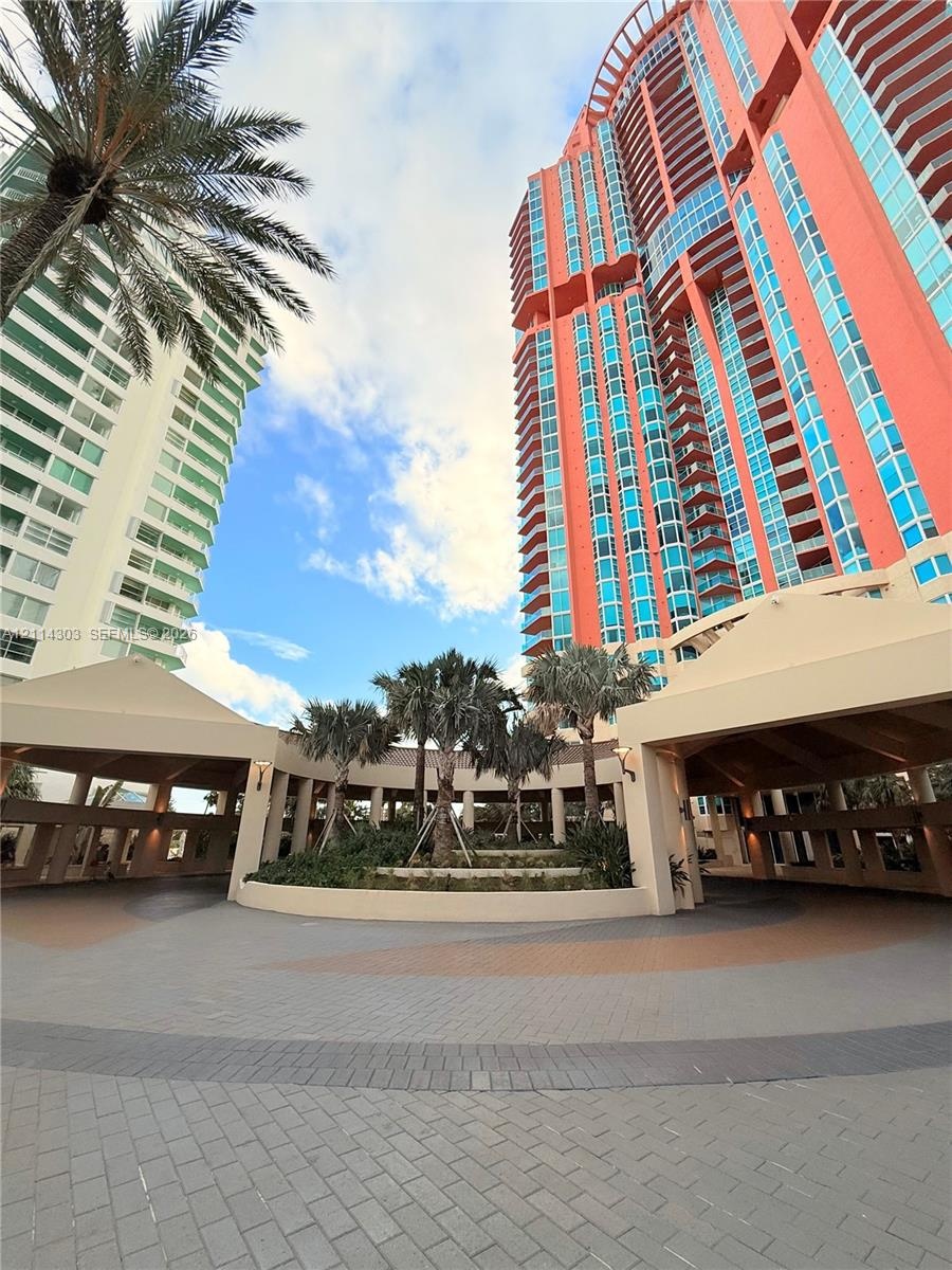 Multi-story high-rise building with a glass facade, circular paver driveway, covered porte-cochère, and a central landscaped courtyard featuring matur