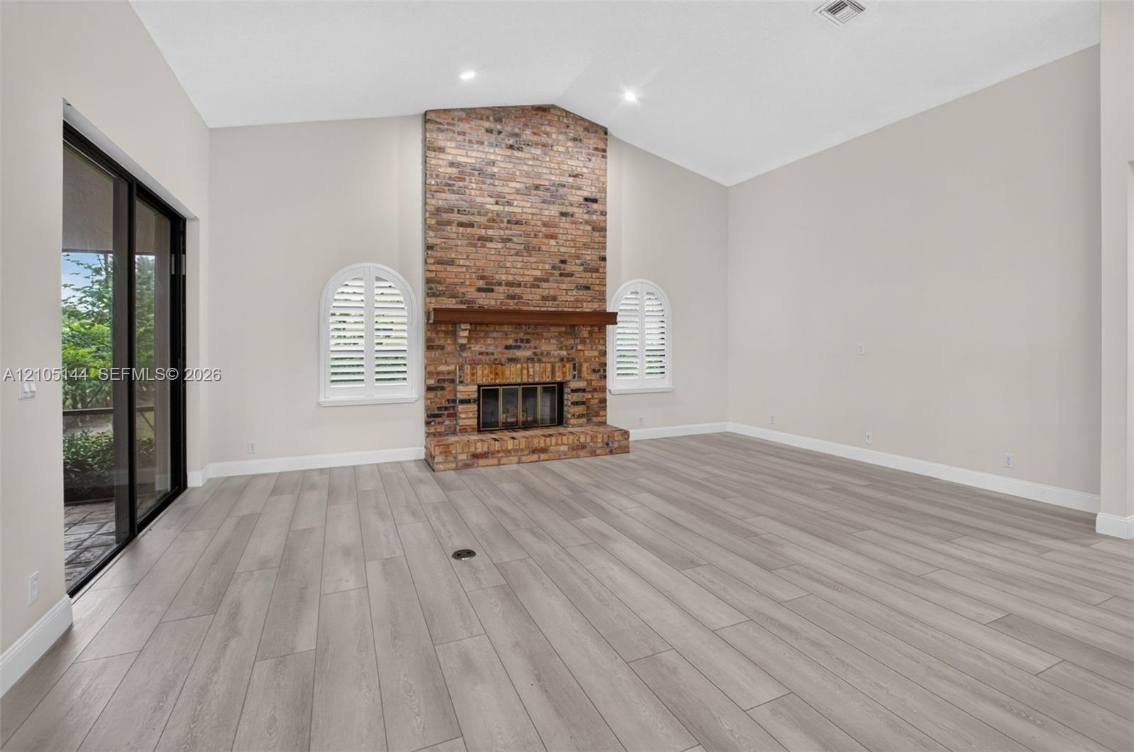 Vaulted ceiling, floor-to-ceiling brick fireplace with wood mantel, wood-finish flooring, arched windows with plantation shutters, and sliding glass d