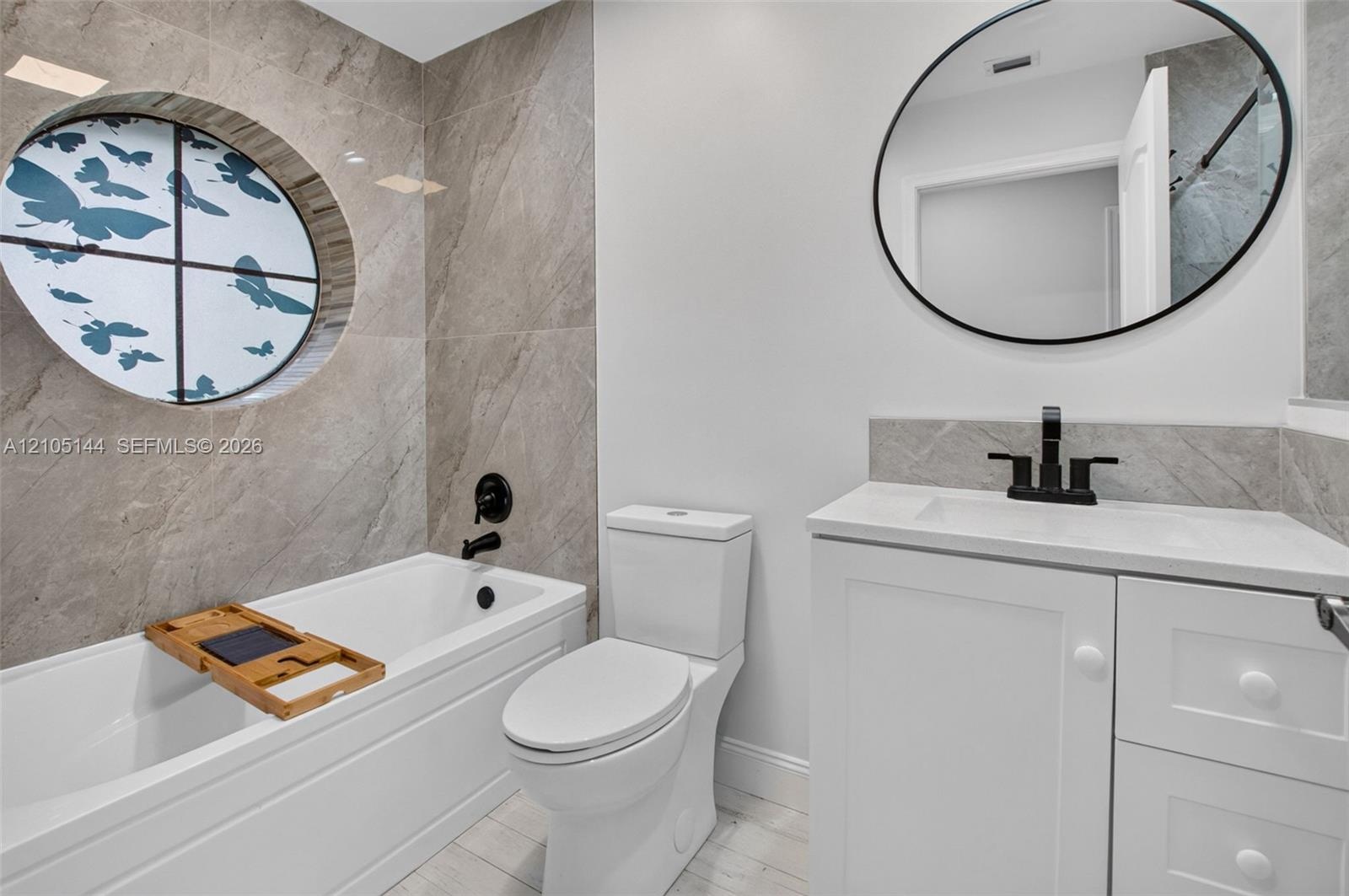 Bathroom featuring large-format tile surround, circular accent window, white vanity with integrated basin, matte black fixtures, and oval wall-mounted