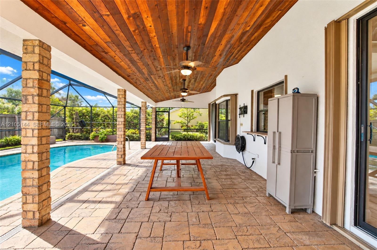 Covered outdoor patio featuring a wood-plank ceiling, recessed ceiling fans, brick-clad support columns, paver flooring, and an enclosed swimming pool