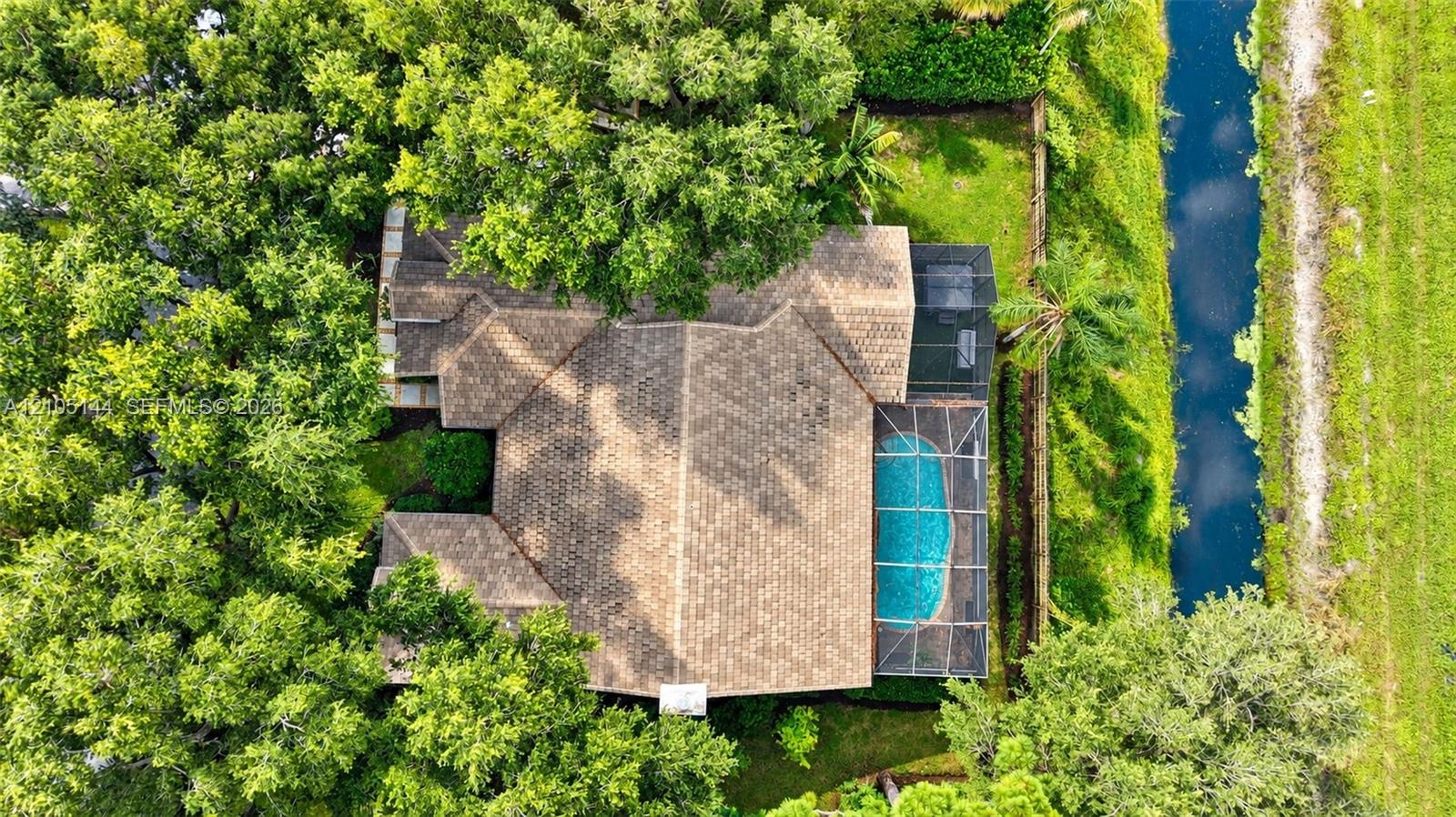 Aerial perspective of a property featuring a tile roof, an enclosed swimming pool, mature tree canopy, and a bordered canal