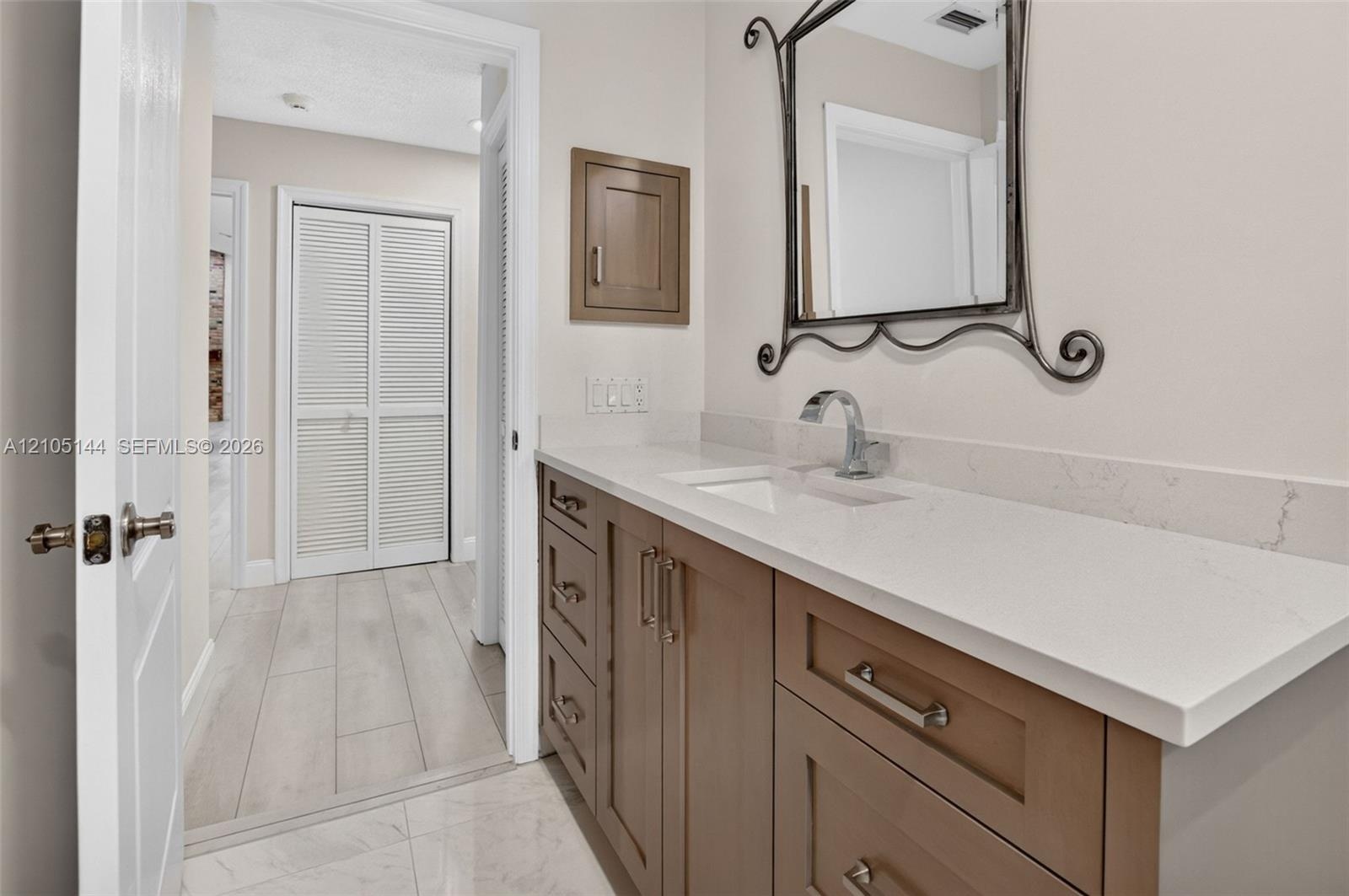 Vanity featuring a stone-composite countertop, undermount sink, shaker-style cabinetry with polished hardware, wall-mounted metallic mirror, and built