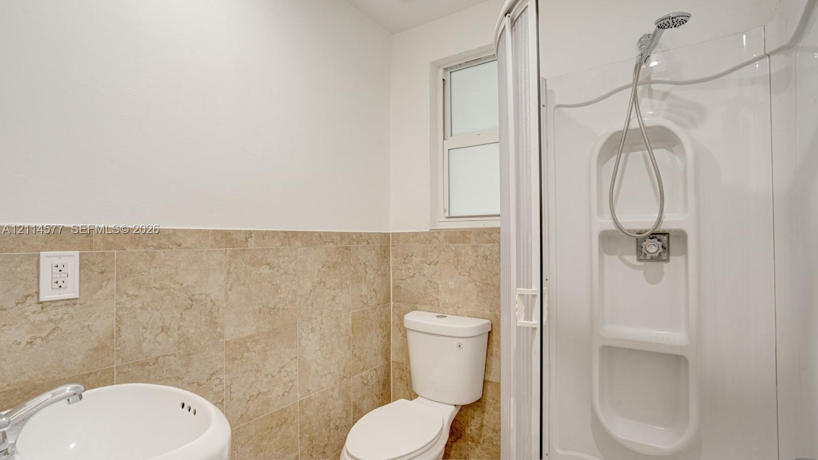 Bathroom featuring large-format beige tile wainscoting, rounded wall-mount basin, white toilet, and enclosed shower stall with built-in shelving