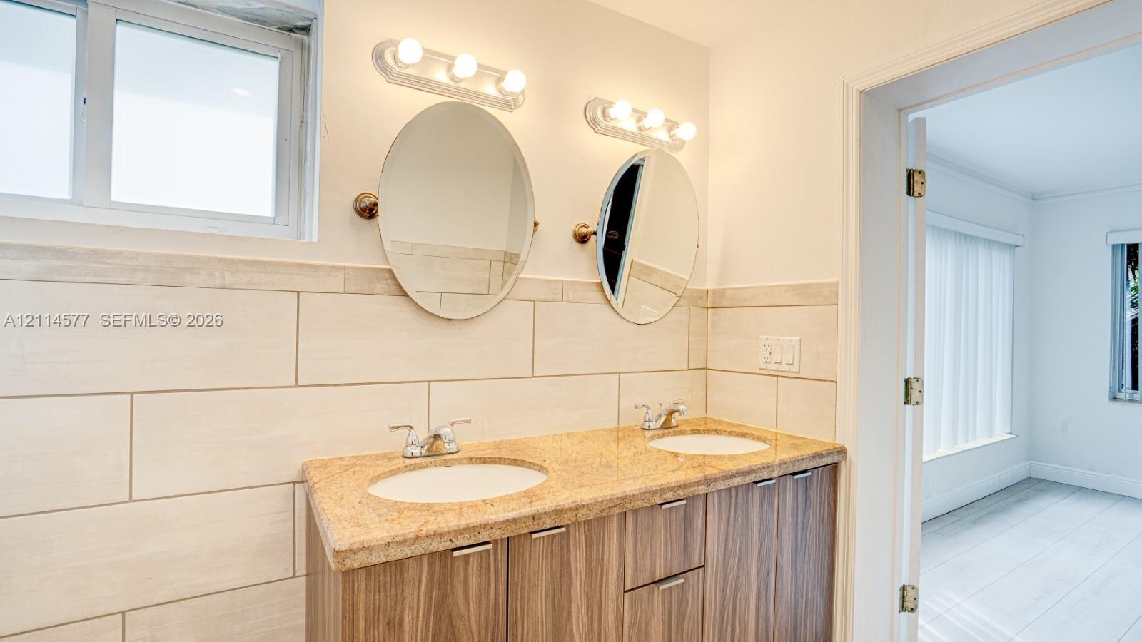 Double vanity with a stone-finish countertop, dual drop-in sinks, wood-finish cabinetry, and ceramic tile wall accents