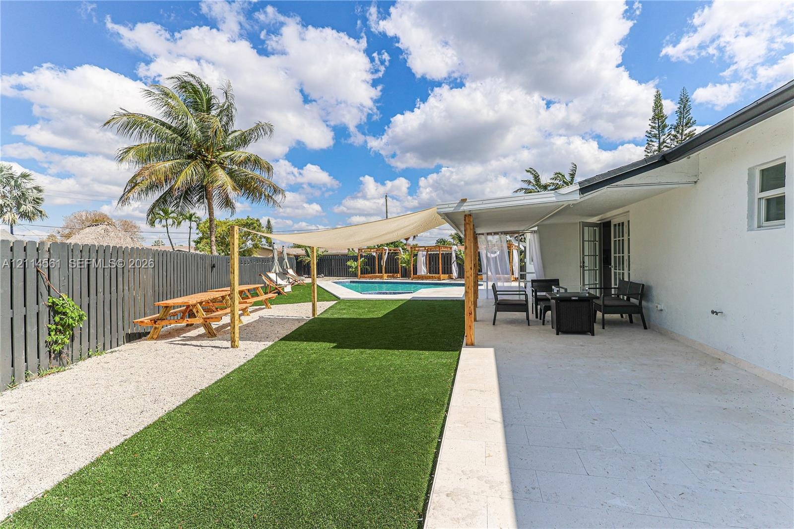 Backyard featuring a paved stone patio, artificial turf lawn, swimming pool, tension shade sail, and perimeter wood privacy fencing