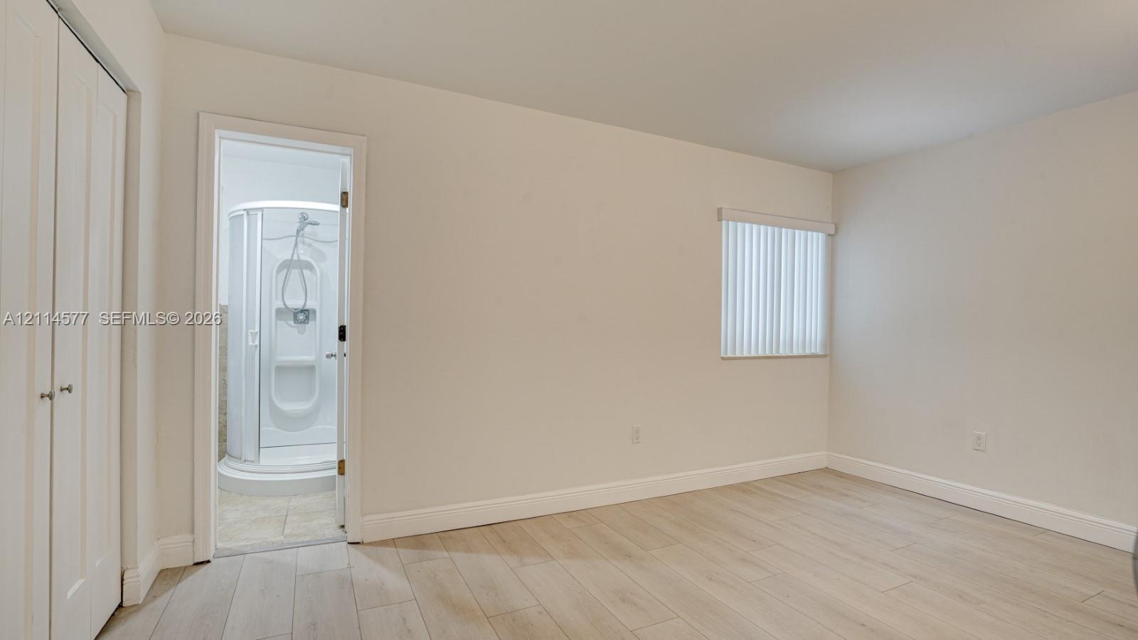 Room featuring wood-finish flooring, white double-door closet, and window with vertical blinds, accompanied by an enclosed shower stall with integrate