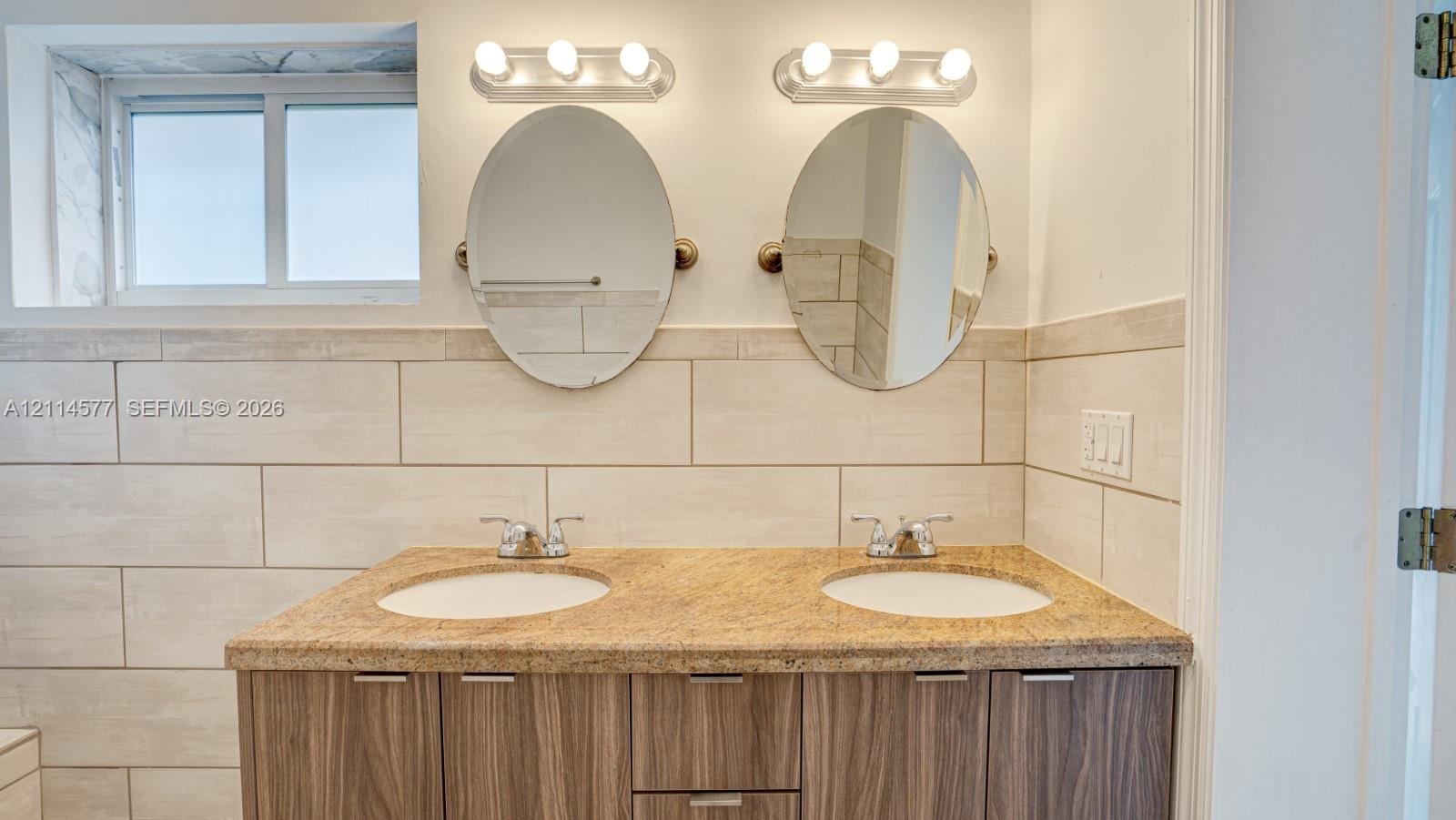 Double vanity featuring a stone countertop, dual undermount sinks, wood-finish cabinetry, two oval mirrors, and horizontal tile wall surrounds