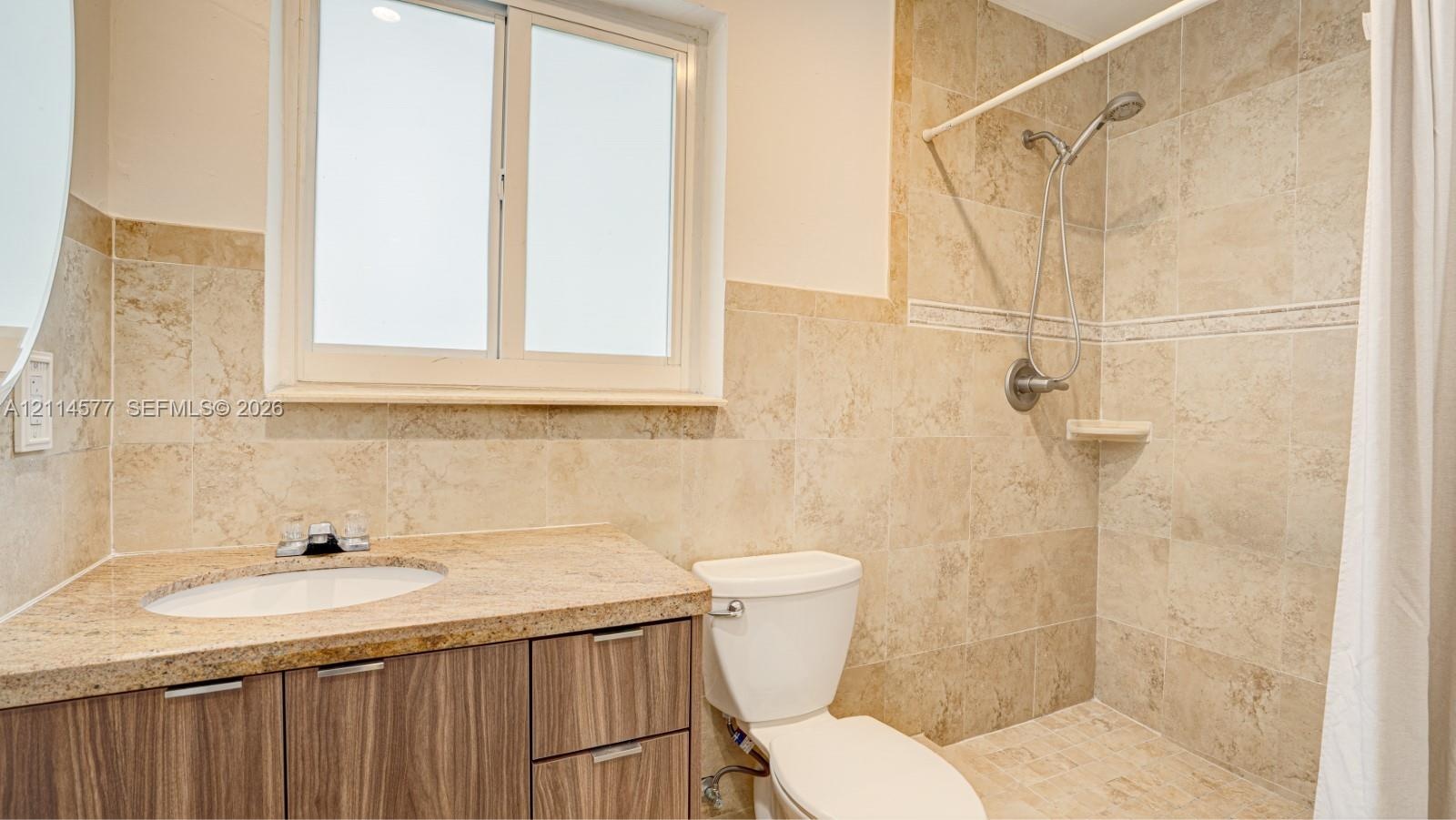 Bathroom featuring stone-tile wall surround, wood-finish vanity cabinetry, stone-slab countertop with integrated oval sink, and a handheld shower fixt