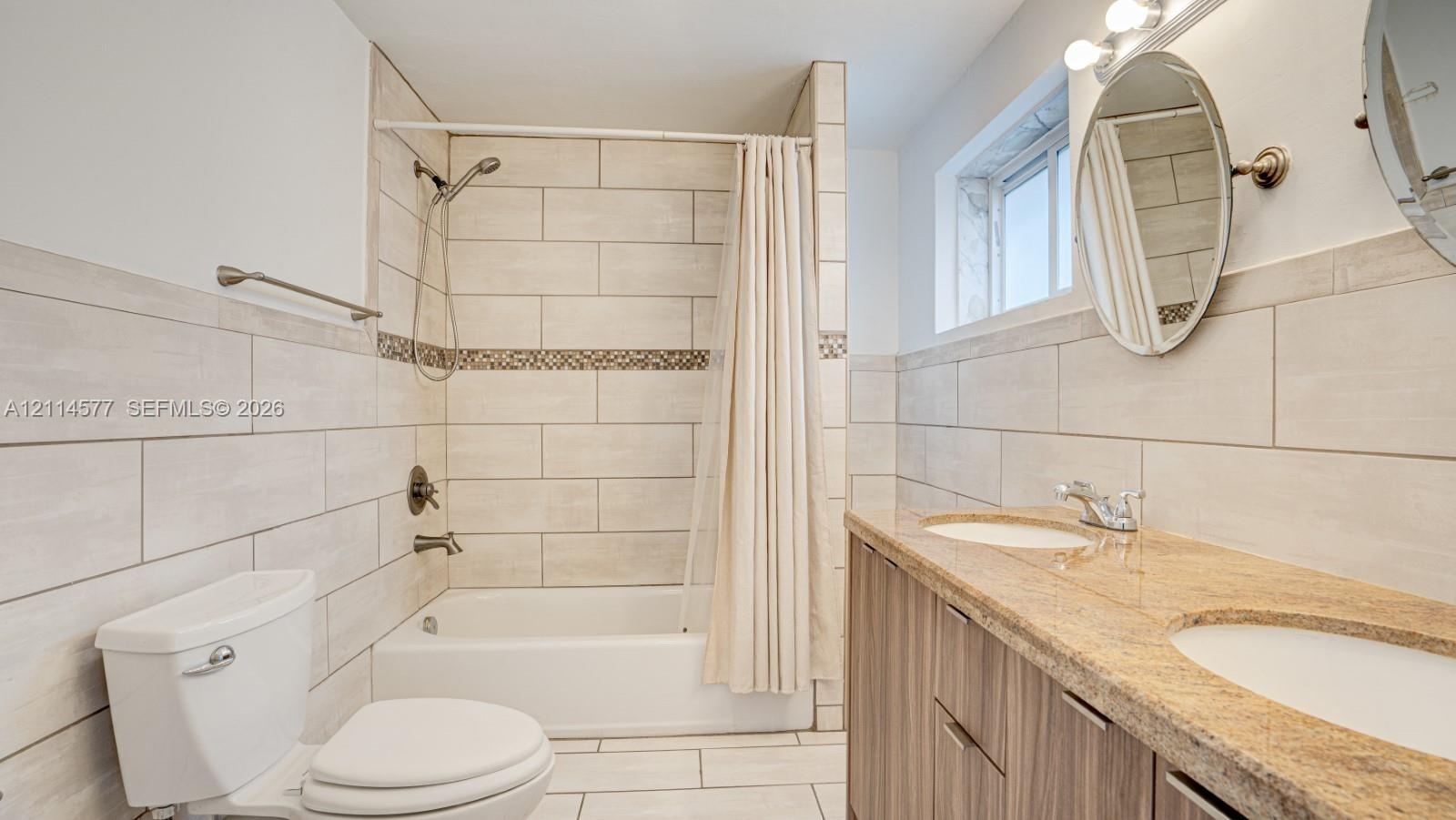 Dual-sink vanity with stone countertop, large-format tile surround, bathtub and shower combination, and oval wall-mounted mirrors