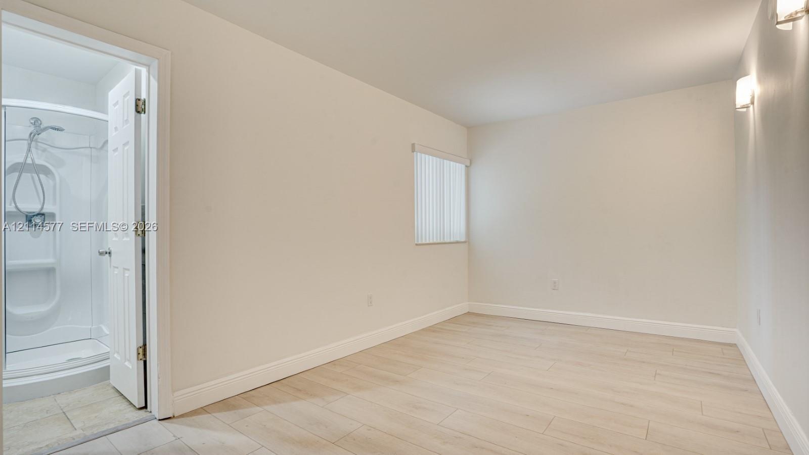 Neutral-toned room with wood-finish flooring, a vertical blind window covering, wall-mounted light fixtures, and a shower stall enclosure with a handh