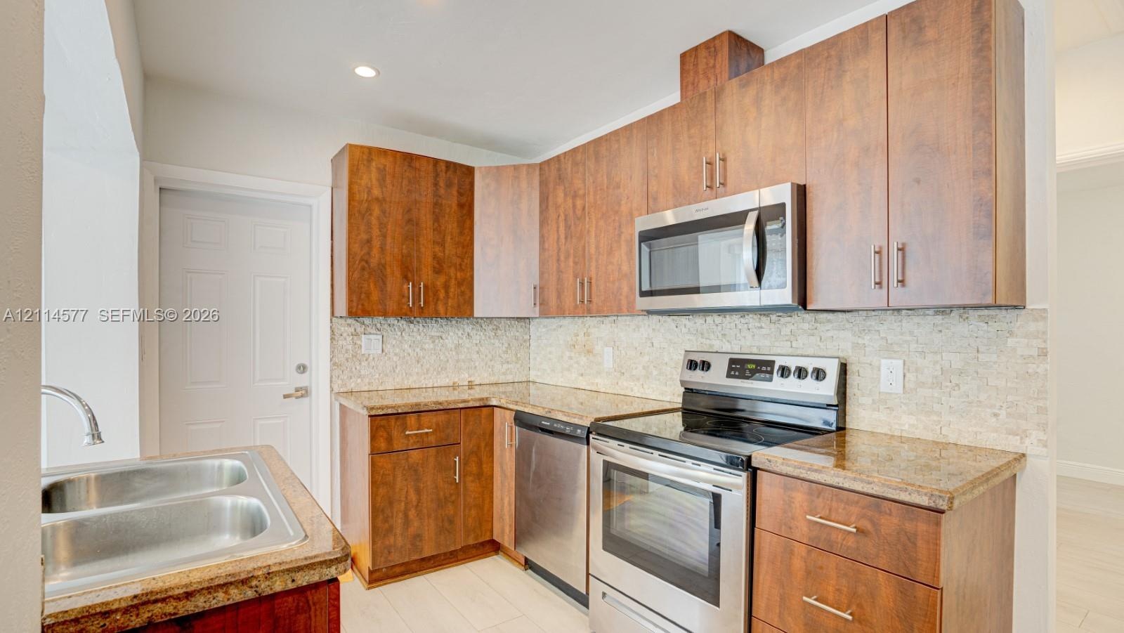 Kitchen featuring flat-panel wood-finish cabinetry, stone-surface countertops, textured stone backsplash, stainless steel appliances, and a double-bas