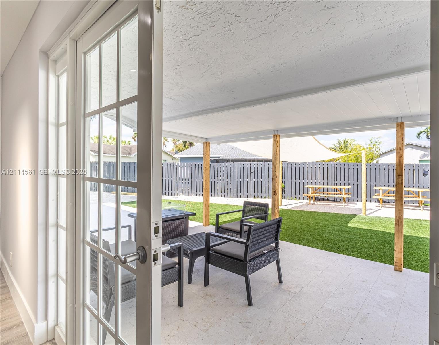 Covered patio with light-toned stone tile flooring, wooden support columns, white-framed glass doors, artificial turf, and a perimeter fence