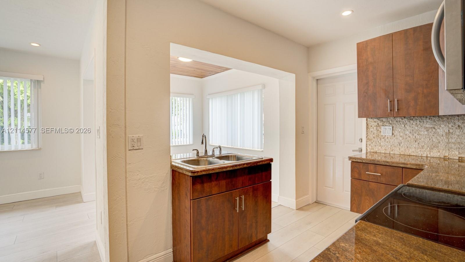 Kitchen featuring wood-finish cabinetry, stone-texture countertops, a dual-basin stainless steel sink, tiled backsplash, and light-toned wood-finish f