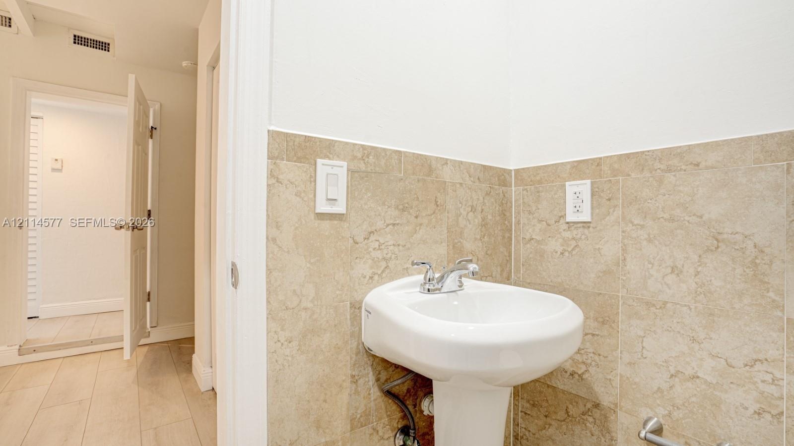 White pedestal sink featuring a dual-handle chrome faucet, complemented by neutral-toned tile wainscoting and light wood-finish flooring